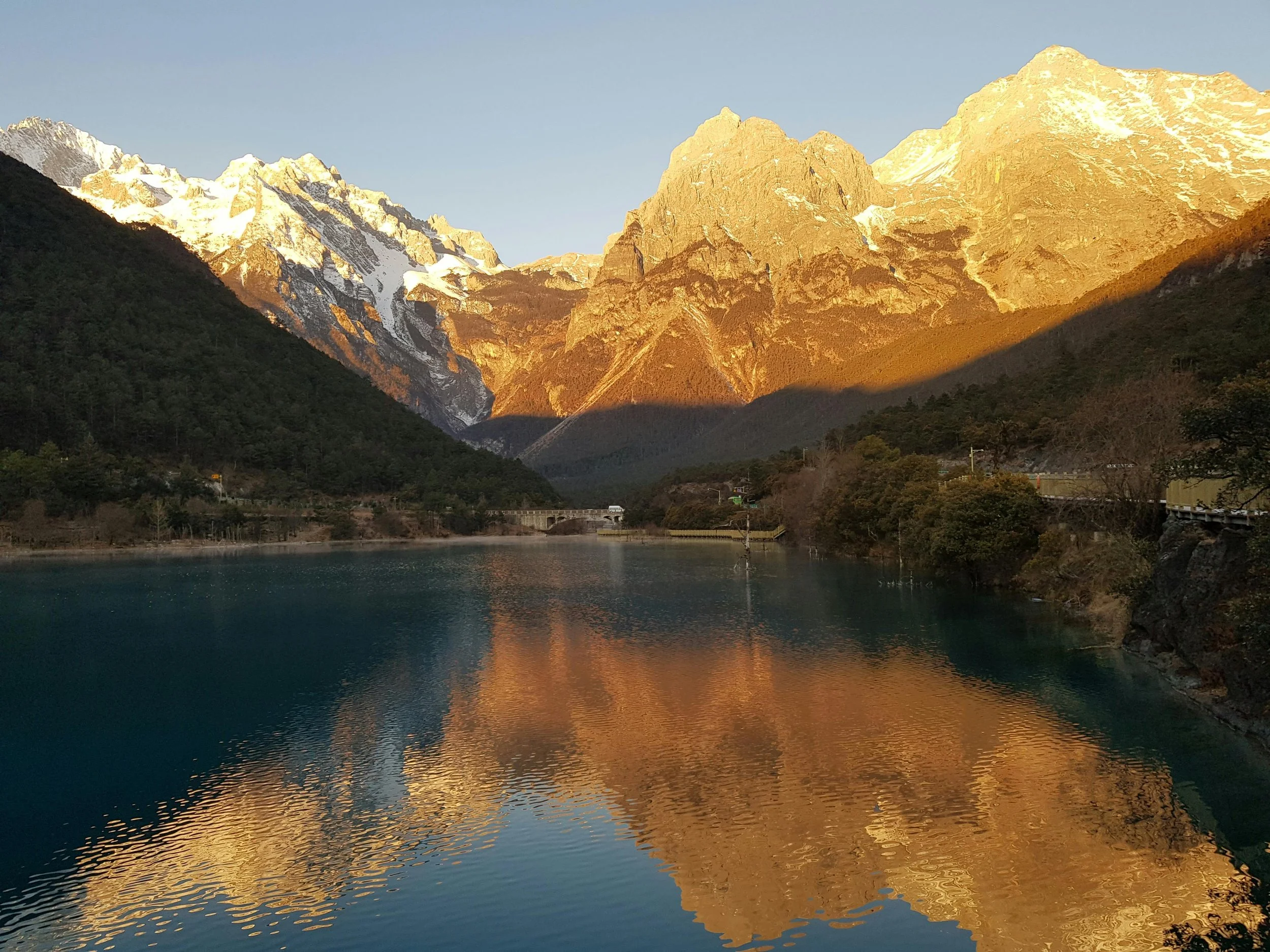 Snow-capped mountains reflecting in a calm lake with forested hills on either side.