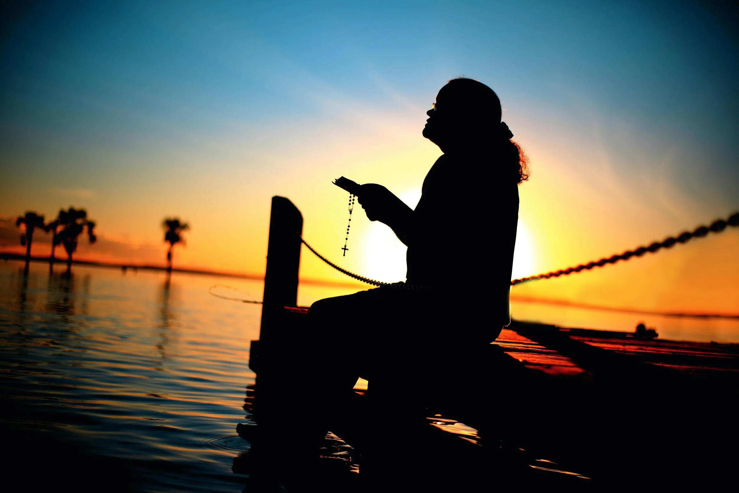 Silhouette of a person sitting on a dock at sunset, holding a rosary and looking at a mobile phone, with palm trees in the background and calm water.
