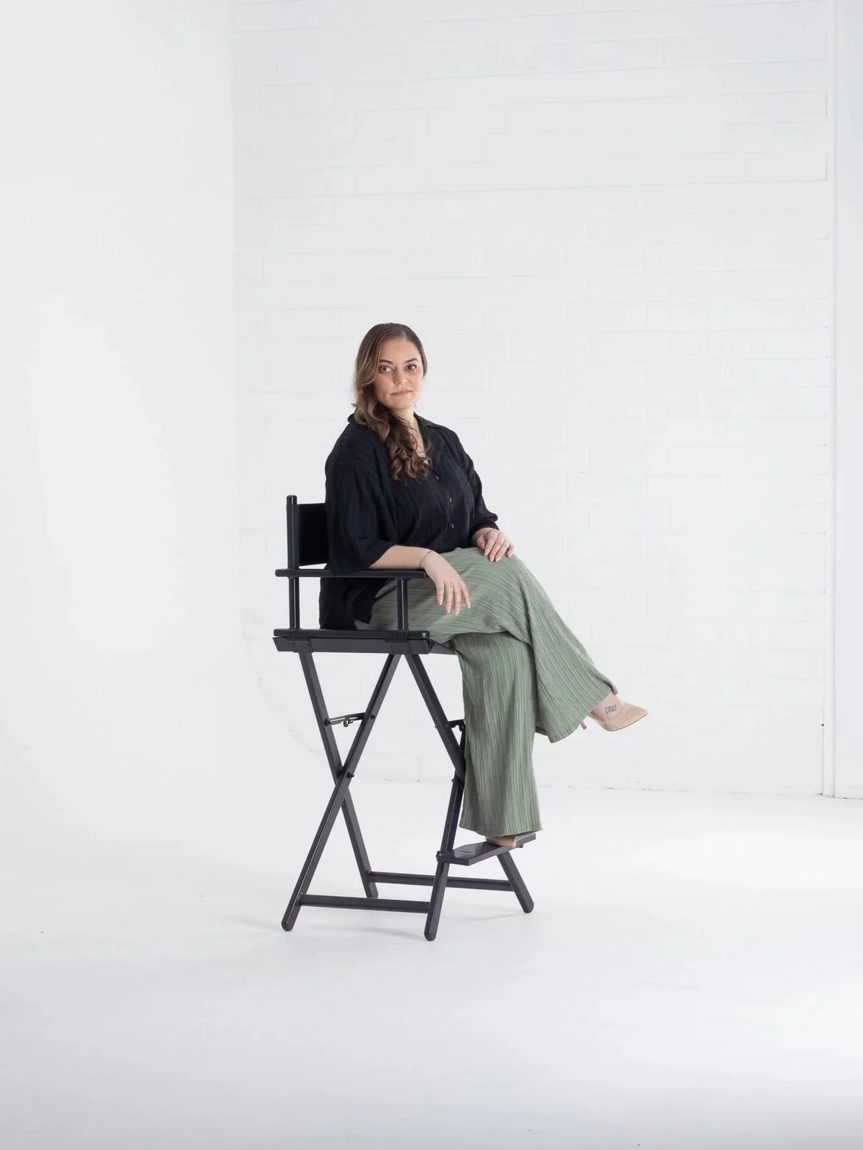 A woman sitting on a high black director's chair in a white studio, wearing a black top and green wide-leg pants.