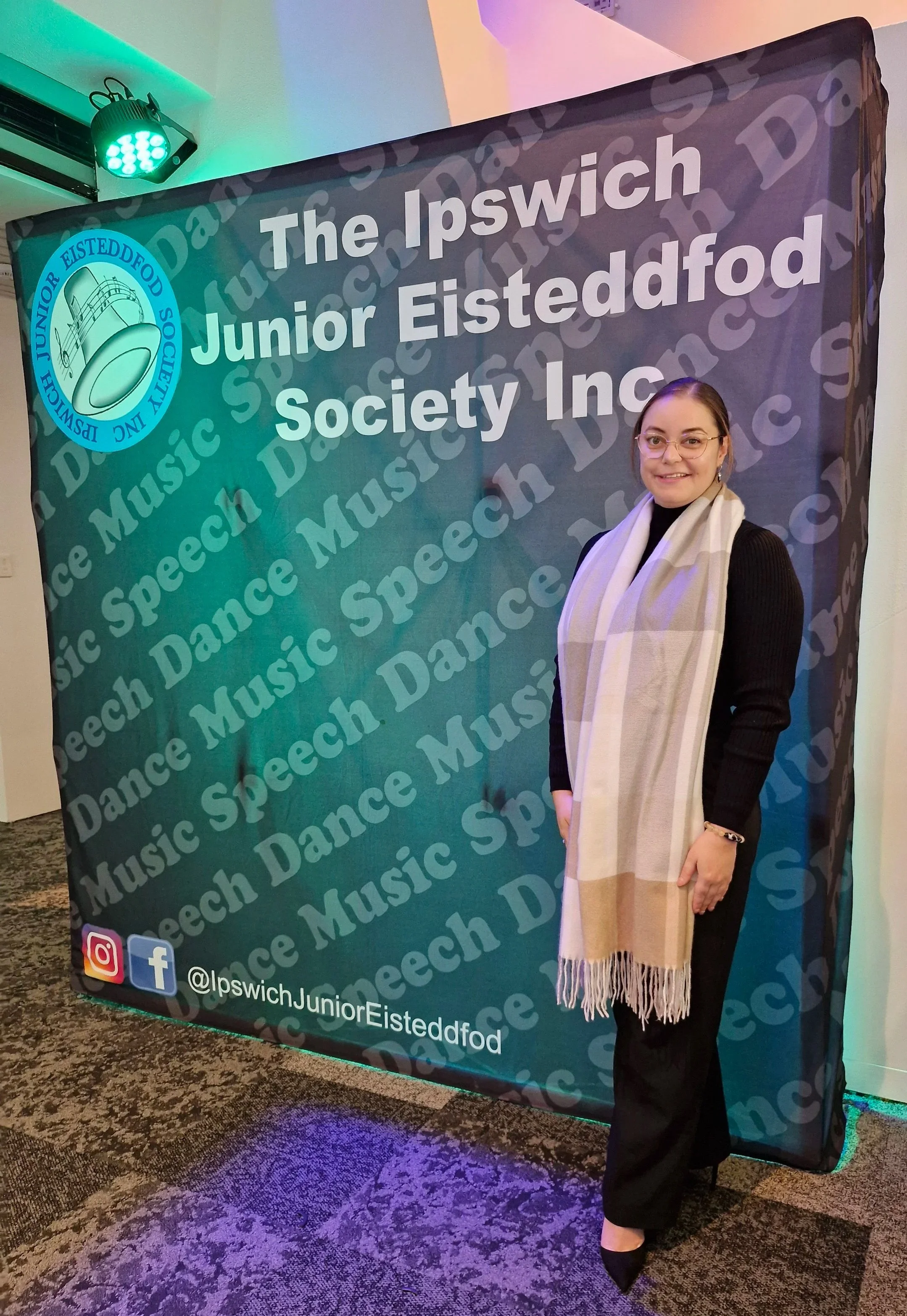 A woman standing in front of a large banner for the Ipswich Junior Eisteddfod Society Inc. with the organization's logo of a bell and musical notes. The banner has a repeating background pattern of the words "Music," "Speech," "Dance," and "Speech Dance." The woman is smiling, wearing glasses, a black outfit, and a large beige plaid scarf.