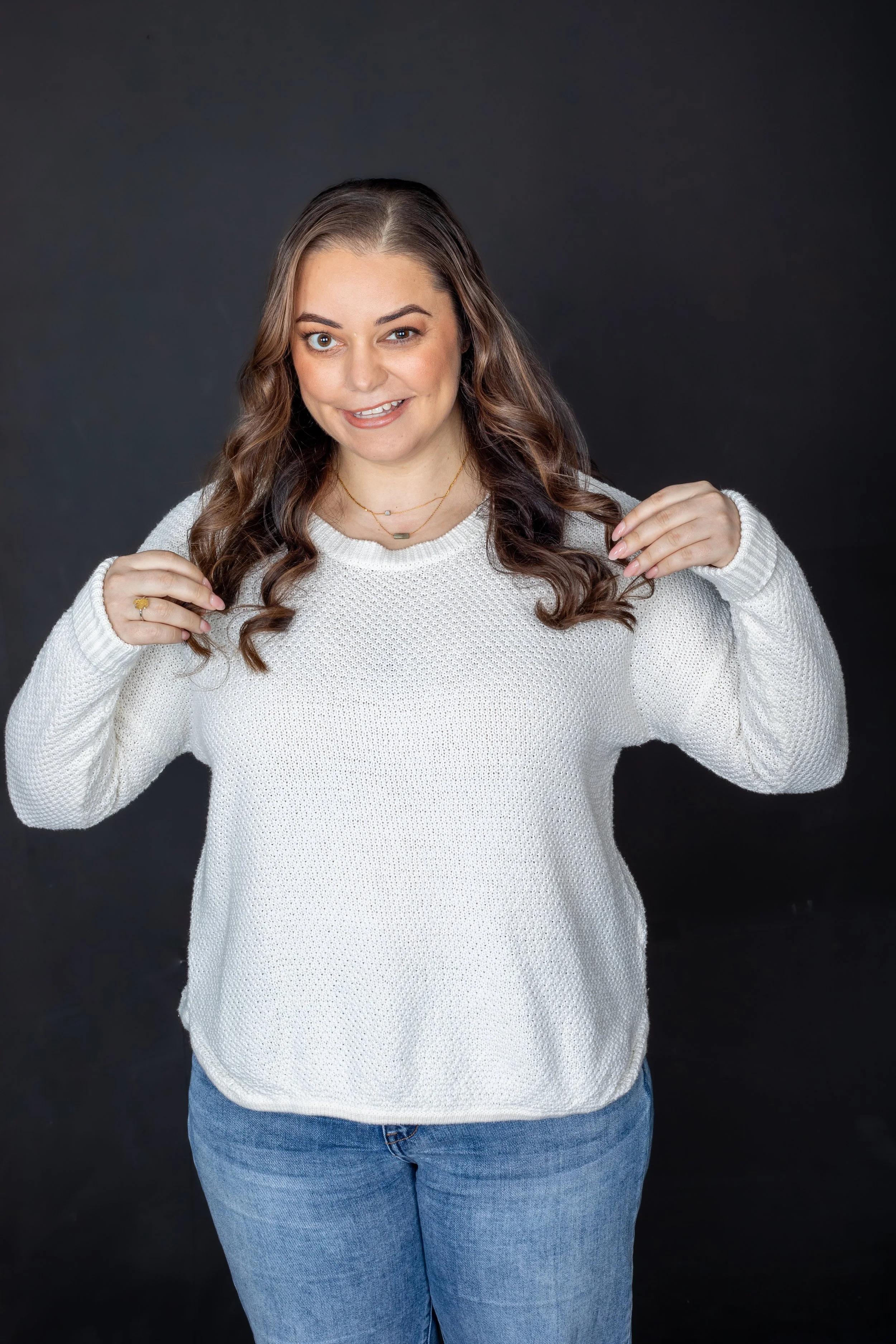 A woman with long wavy brown hair wearing a white knit sweater and blue jeans against a dark background, smiling and holding her hair with both hands.