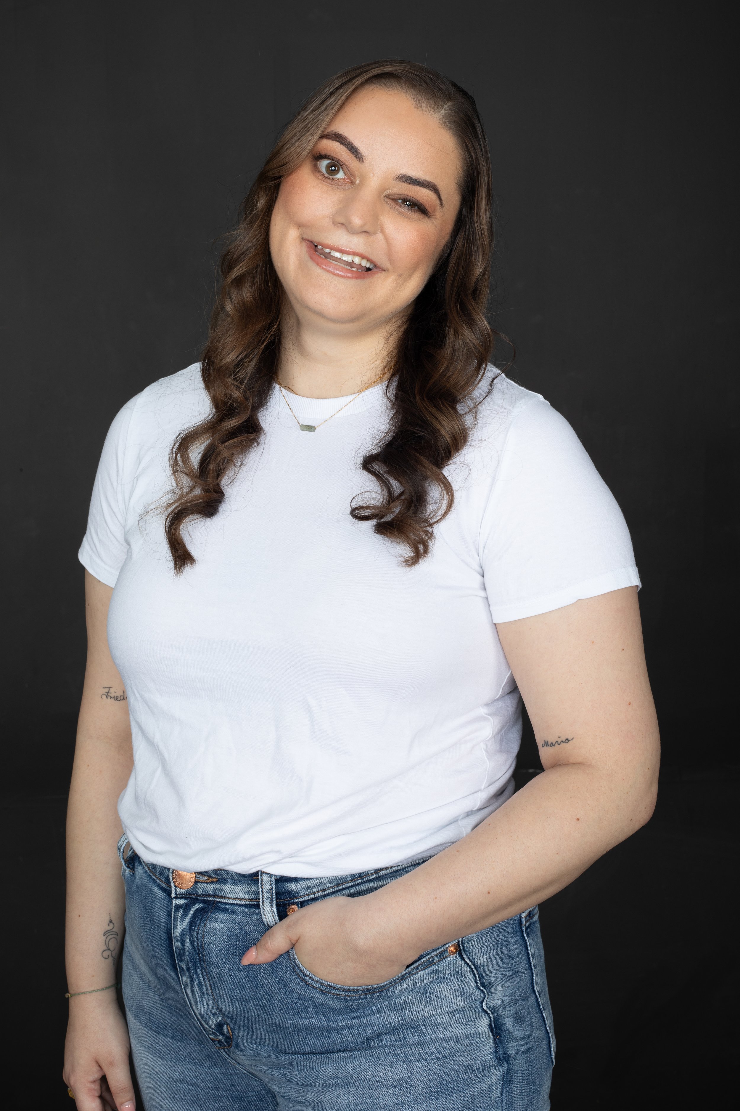 A woman with long brown hair, wearing a white t-shirt and blue jeans, standing against a black background, smiling with one eye closed and her hand in her pocket.