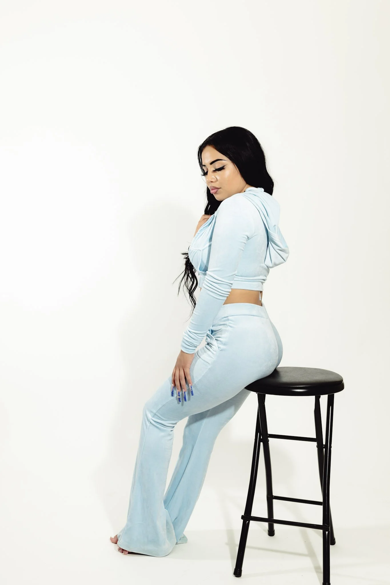 A woman with long black hair, wearing a light blue hoodie and matching pants, is sitting sideways on a black stool against a plain white background. She has her eyes closed and is touching her shoulder with one hand.