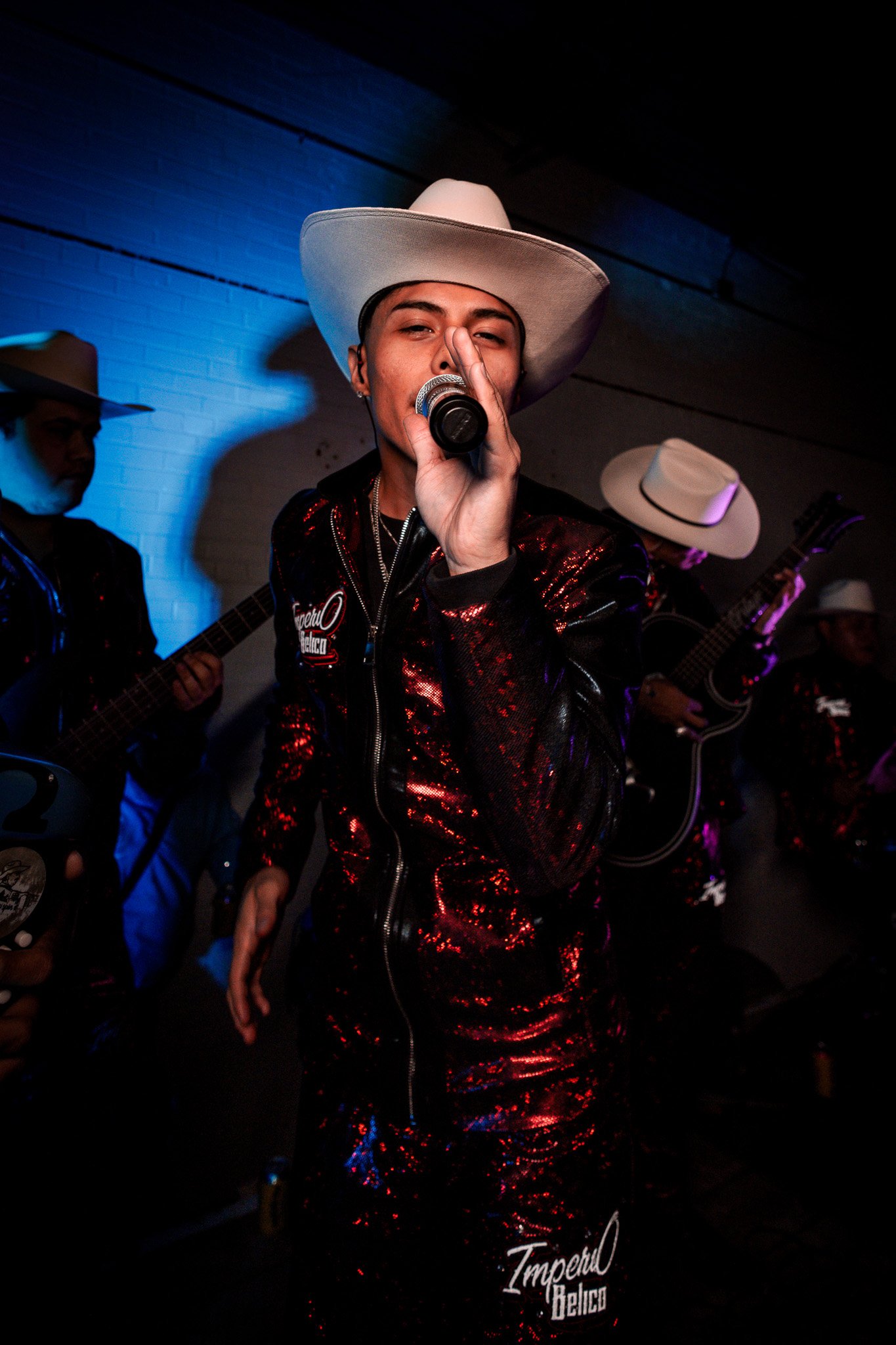 A person singing into a microphone with others playing guitars in the background, wearing a white cowboy hat and a shiny black and red outfit.