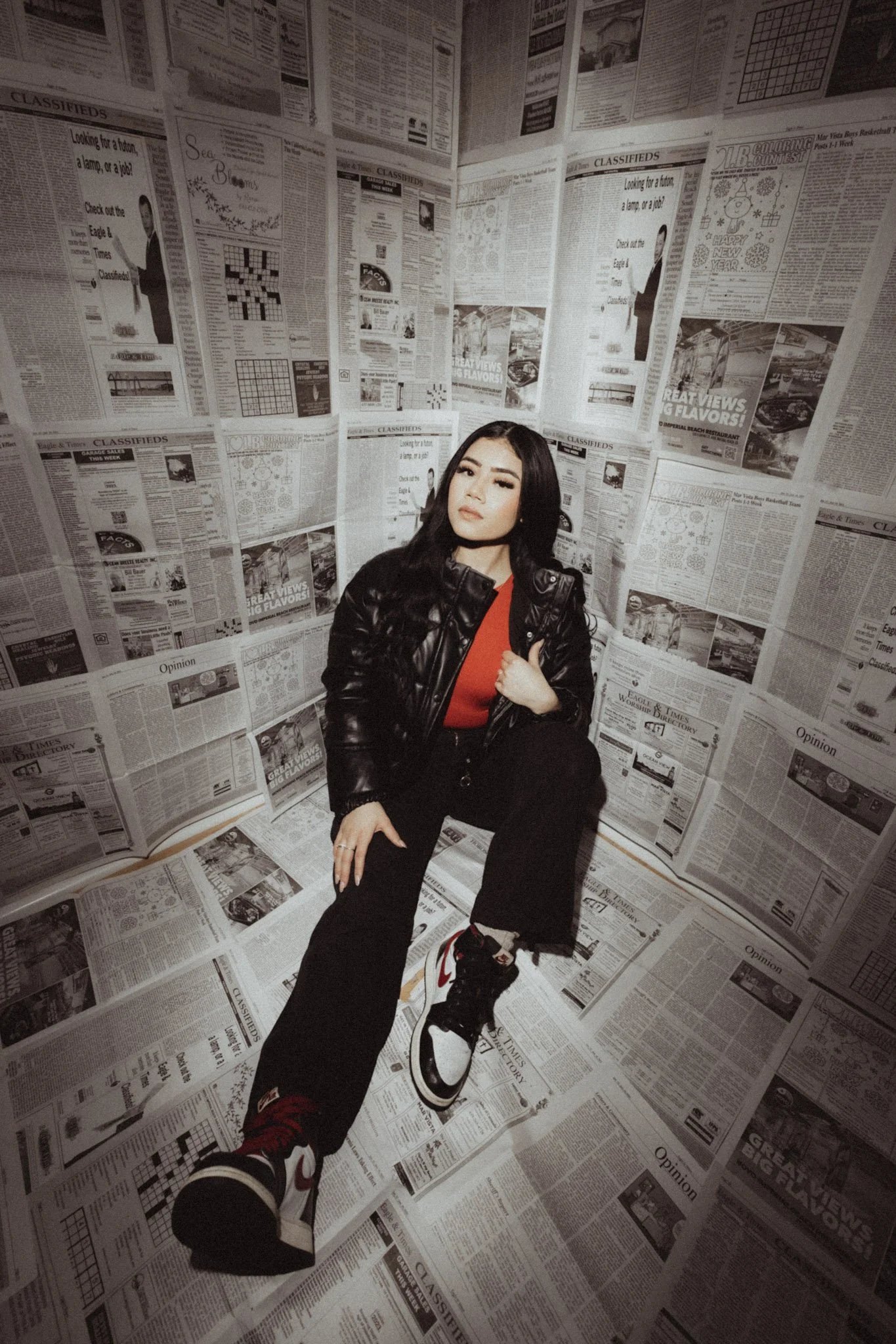 Young woman with long black hair and a leather jacket sitting in a small room with walls and floor covered in newspapers.