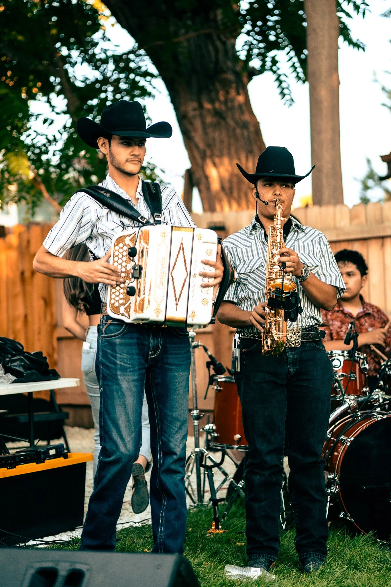 Two musicians in cowboy hats, one playing an accordion and the other playing a saxophone, performing outdoors near a wooden fence and a large tree, with a young player and musical equipment visible.