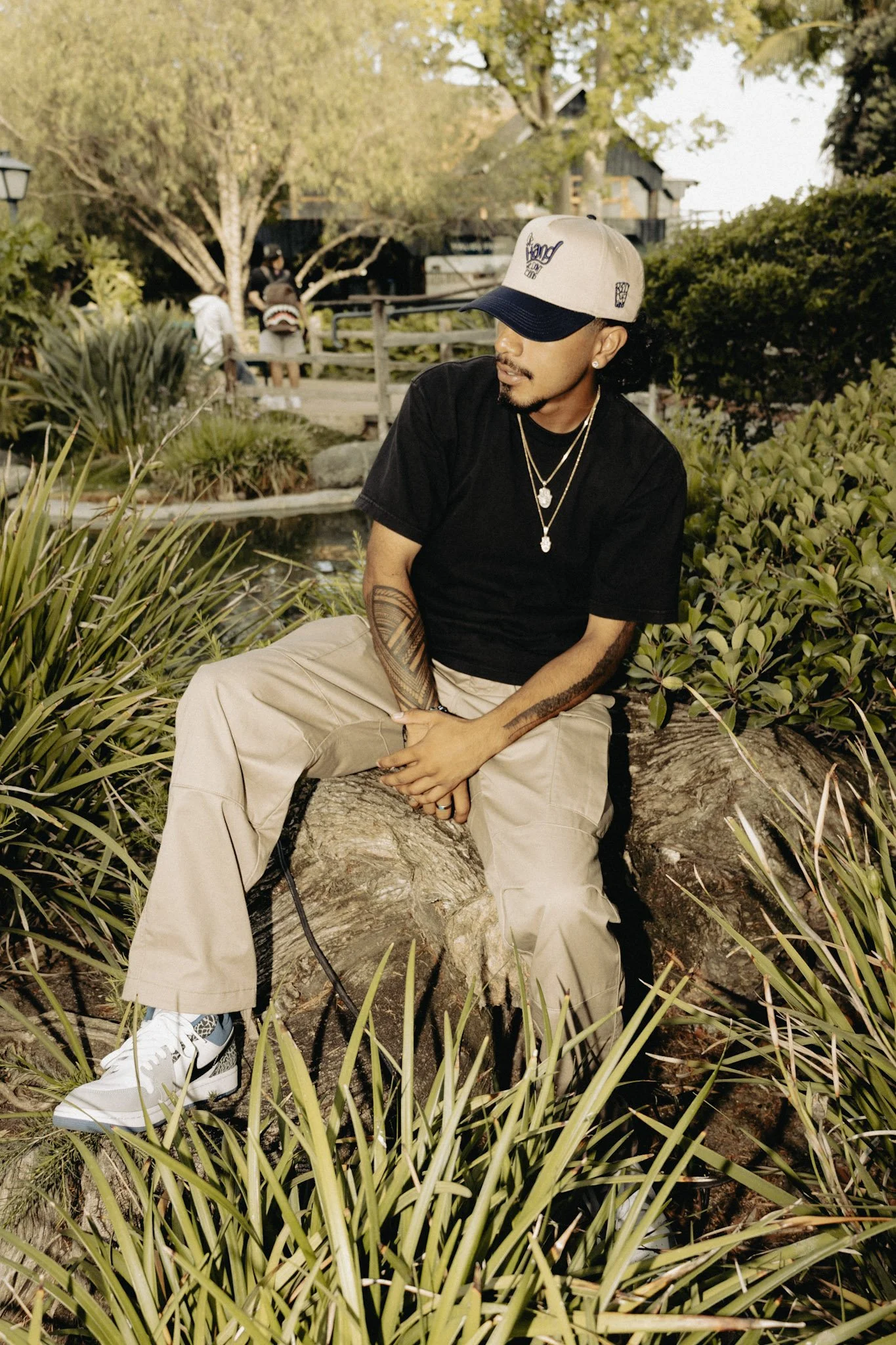 A man sitting on a tree trunk in a park, surrounded by green vegetation and tall grass, wearing a black t-shirt, beige pants, white sneakers, and a cap, with jewelry and tattoos.