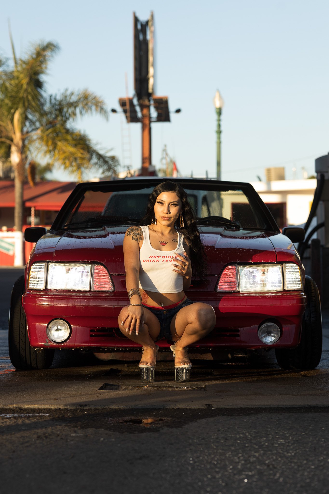 A woman squatting in front of a red sports car during sunset, wearing a white crop top and shorts, with long dark hair and tattoos on her arm.