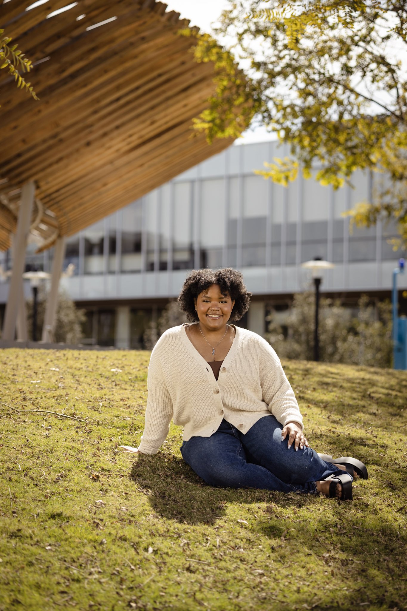 A woman sitting on grass outdoors near a modern building, smiling at the camera. She has curly hair and is dressed casually in a cream cardigan, blue jeans, and black shoes. There are trees with yellowish leaves and a wooden structure overhead.