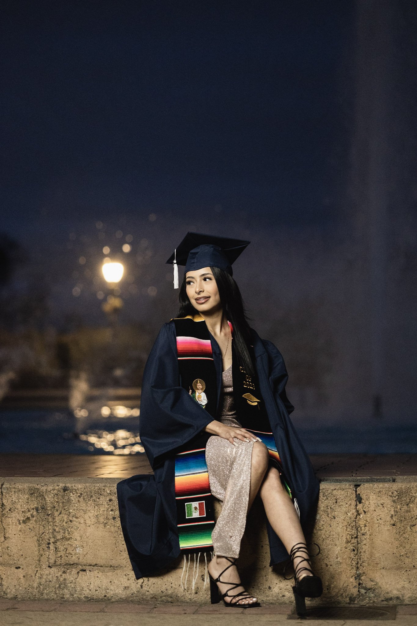 A woman in a graduation cap and gown sitting on a low wall outdoors at night, with a lit streetlamp behind her, smiling and looking off to the side.