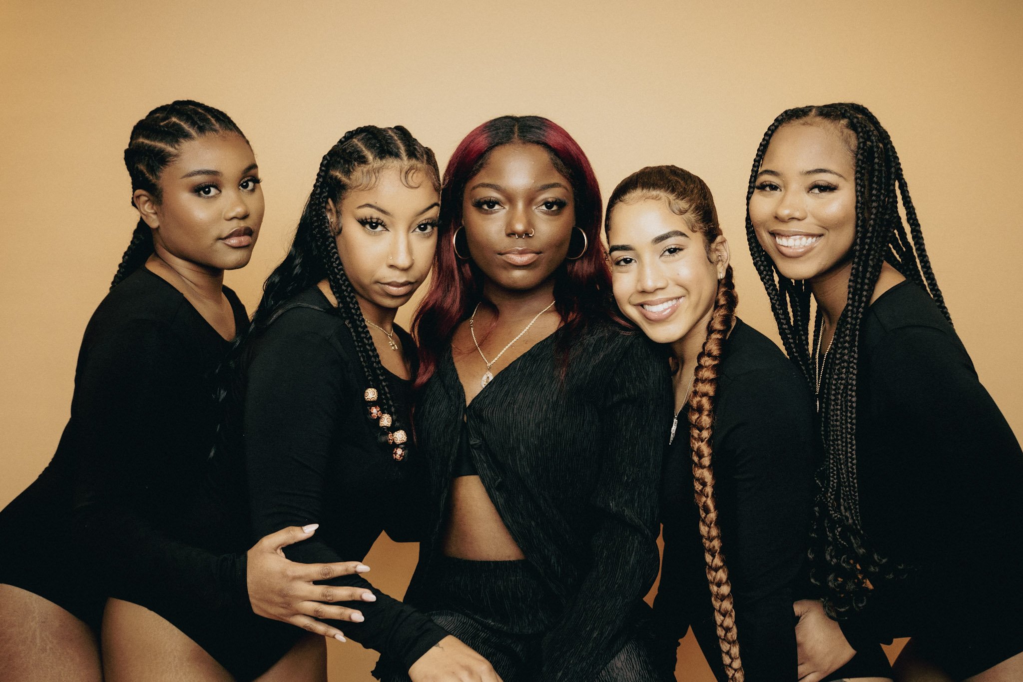Group of five young women posing together against a beige background, wearing black outfits, with various hairstyles and smiles.