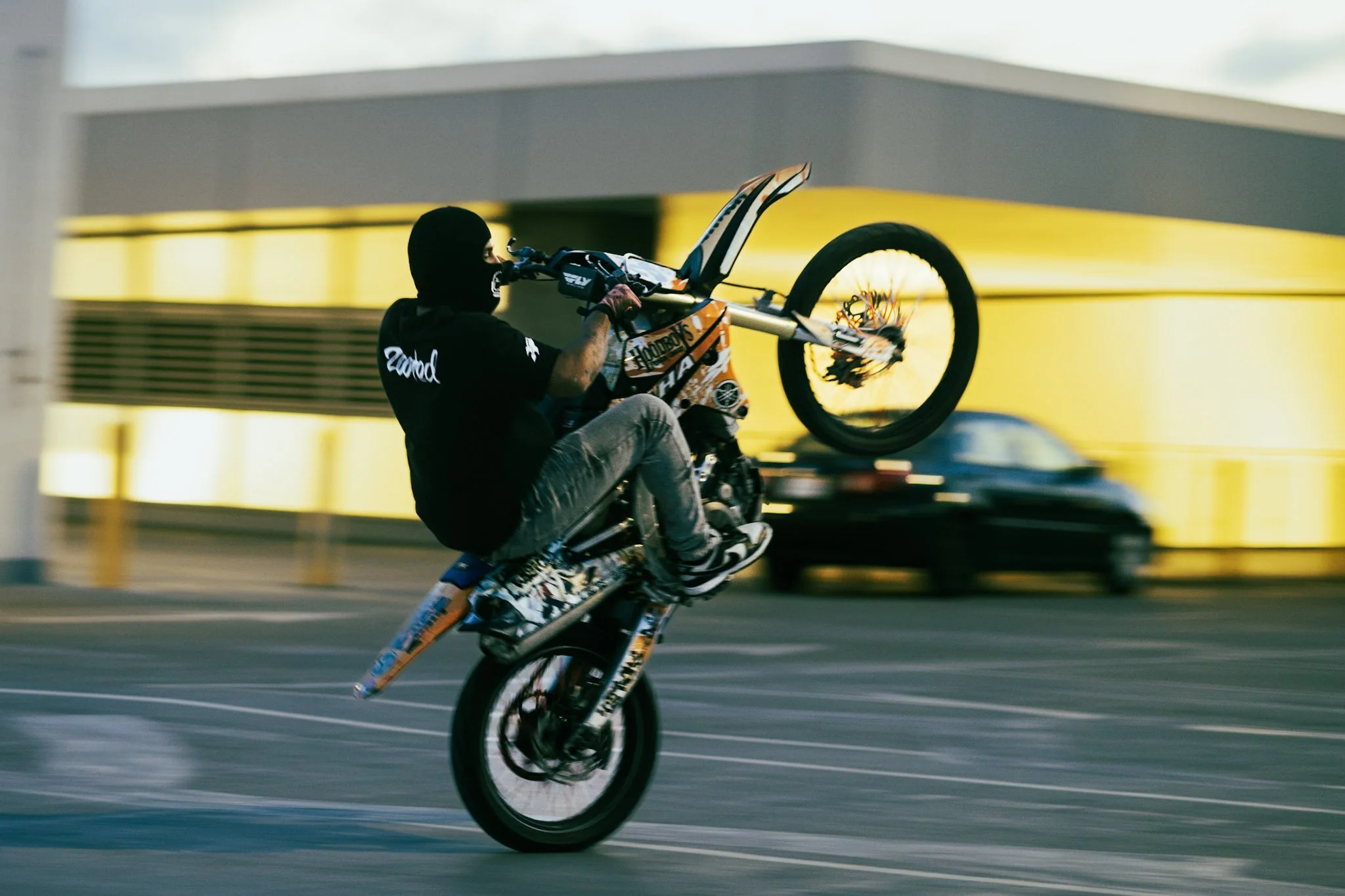 A person wearing a black hoodie and a balaclava performing a stunt on a motocross bike on a city rooftop, with a car passing in the background.