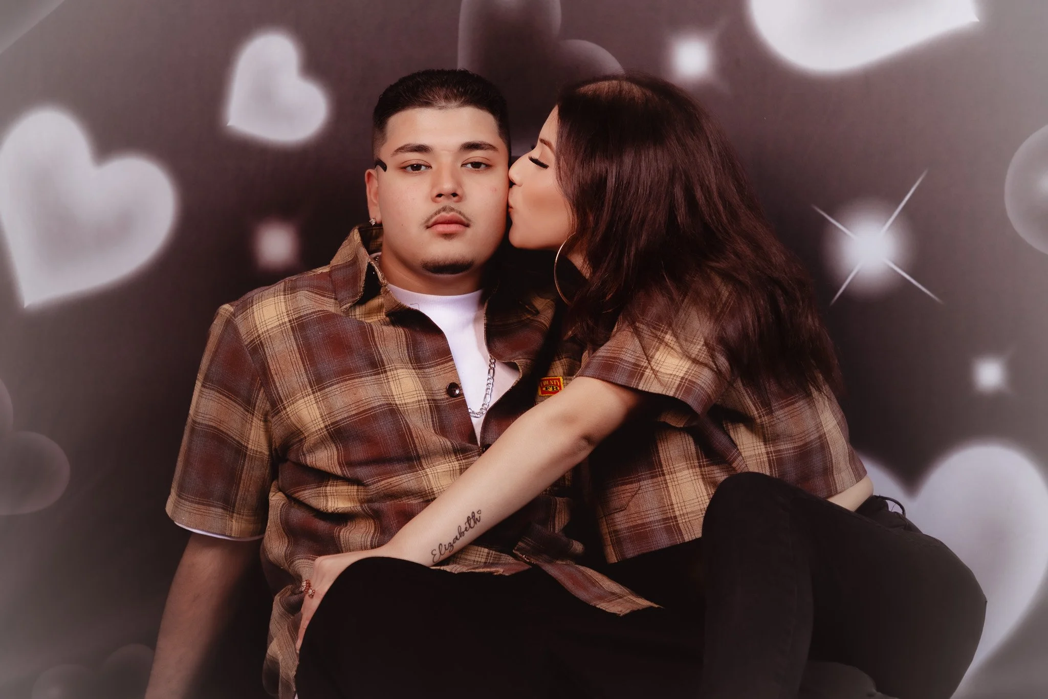 A man with short dark hair and a woman with long dark hair kissing on the cheek, both wearing matching brown plaid shirts, against a backdrop of white hearts and stars.