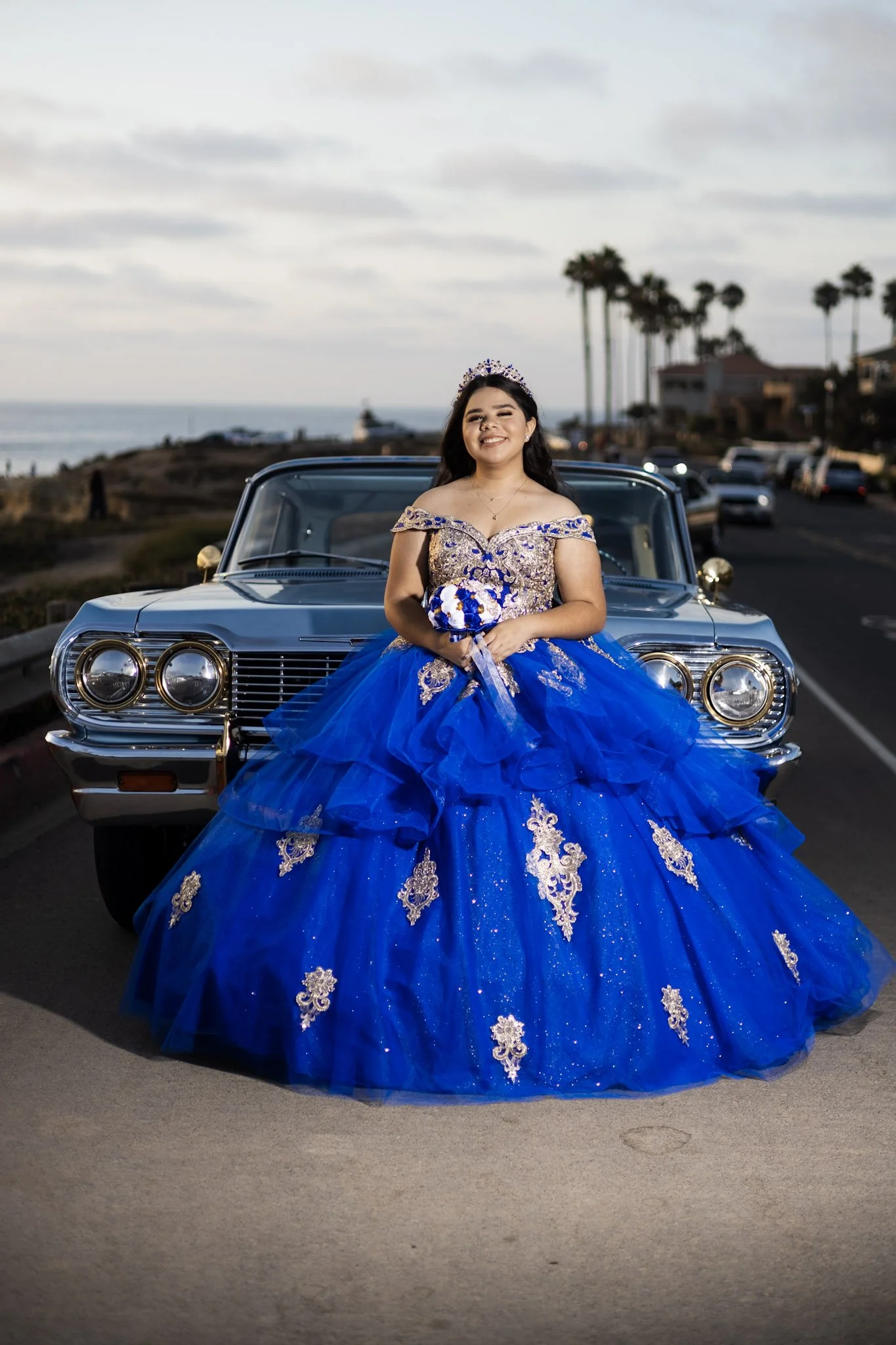 A girl in a royal blue quinceañera dress with gold embroidery and a tiara, holding a bouquet of flowers, standing in front of a vintage car near the coast during sunset.