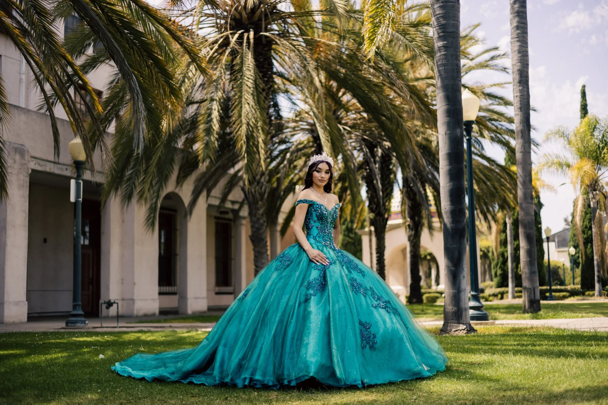 A woman in a turquoise ball gown standing on grass in a park with palm trees and lamp posts, in front of a building.
