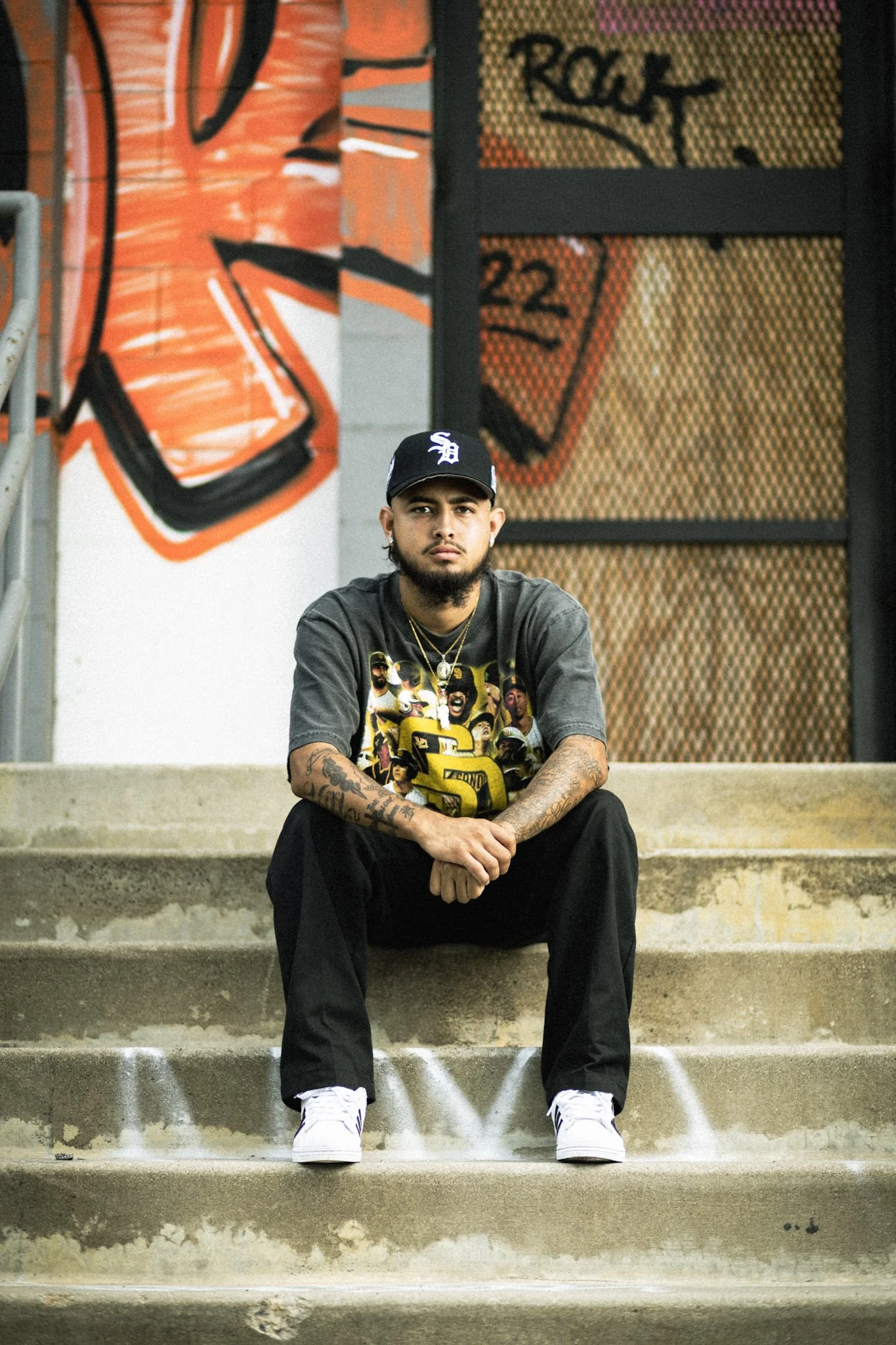 A young man with tattoos, wearing a black cap, gray graphic t-shirt, black pants, and white sneakers, sitting on concrete stairs in front of a colorful graffiti mural featuring the words 'ROCK' and '22'.