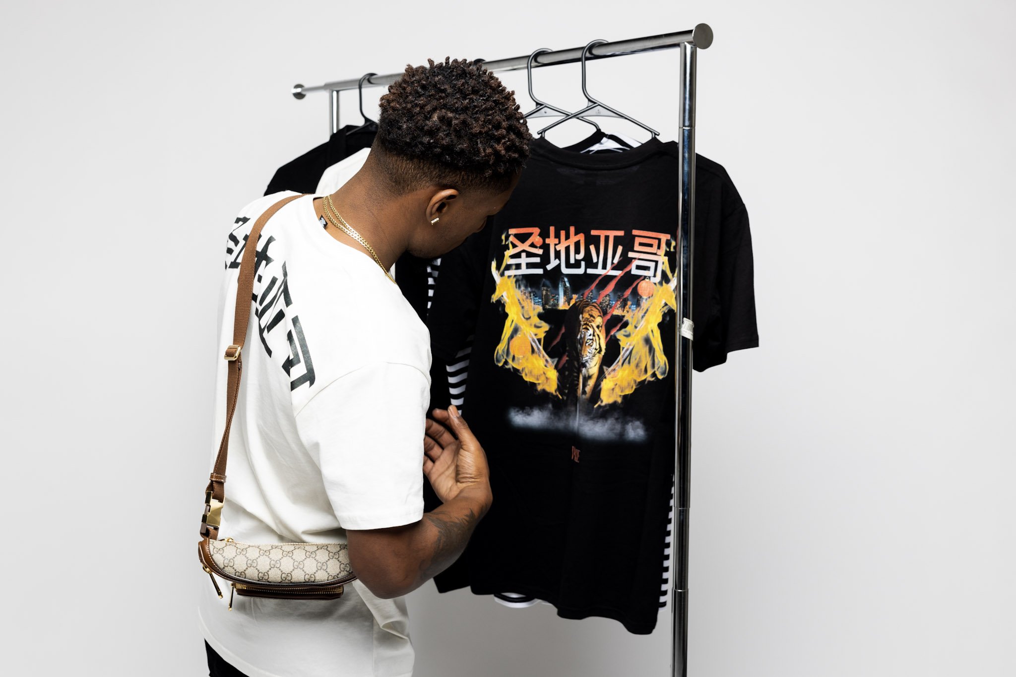 A man with tattoos, wearing a white t-shirt and gold jewelry, is looking at graphic t-shirts hanging on a clothing rack in a minimalist store.