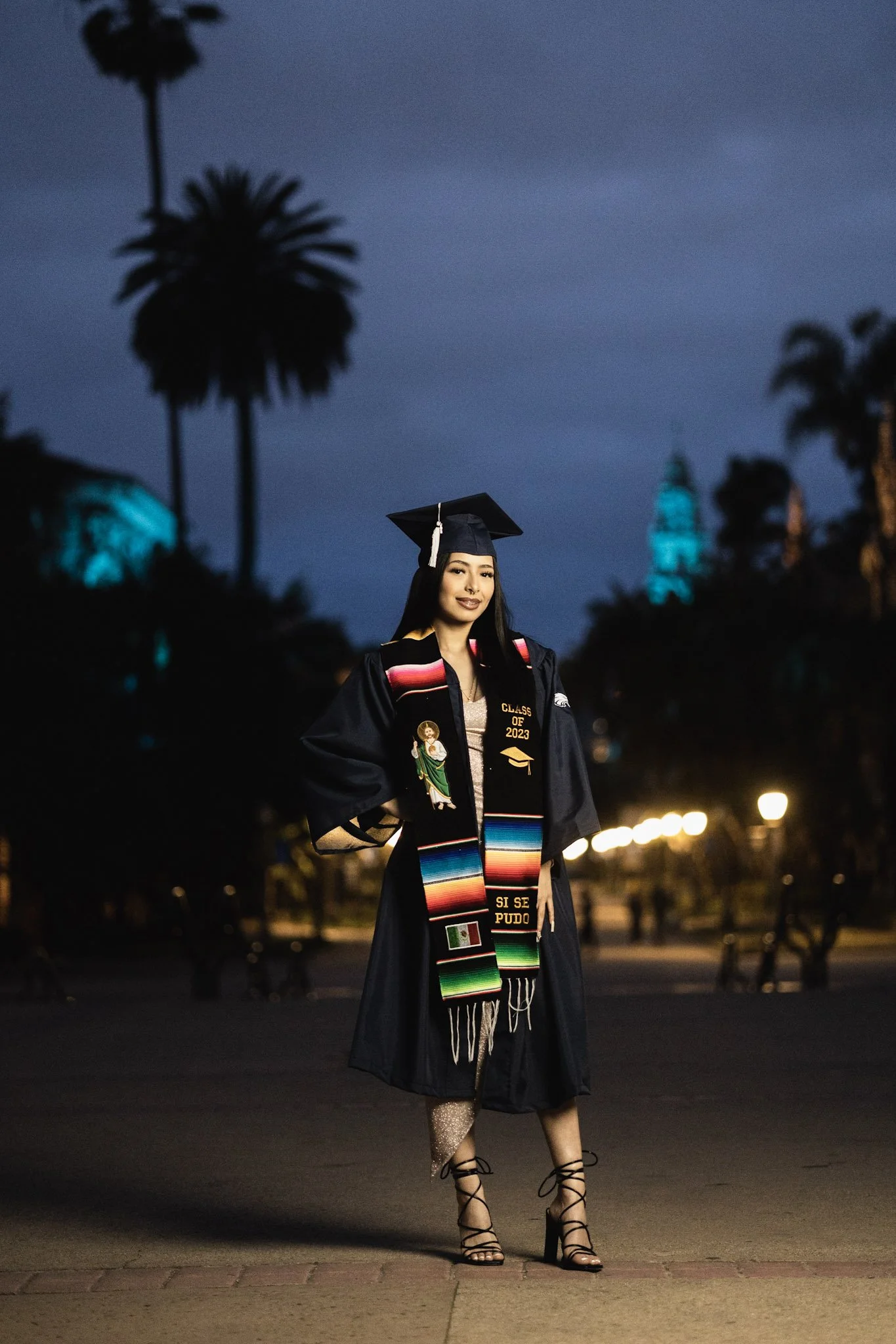 A young woman in a graduation cap and gown standing outdoors at night, smiling, with city lights and palm trees in the background.