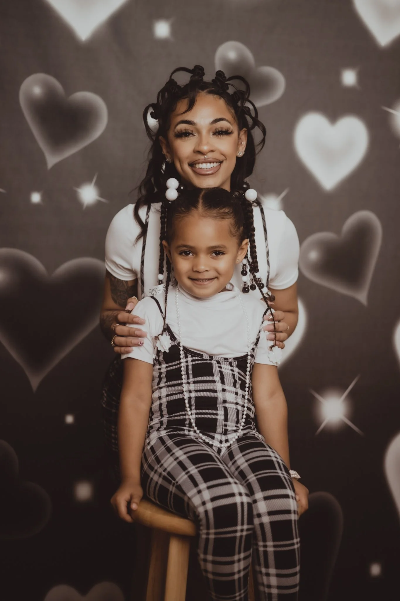 A smiling woman and girl sitting on a stool with a backdrop of hearts and sparkle effects, both wearing white tops and patterned pants or dresses, with the woman gently holding the girl.