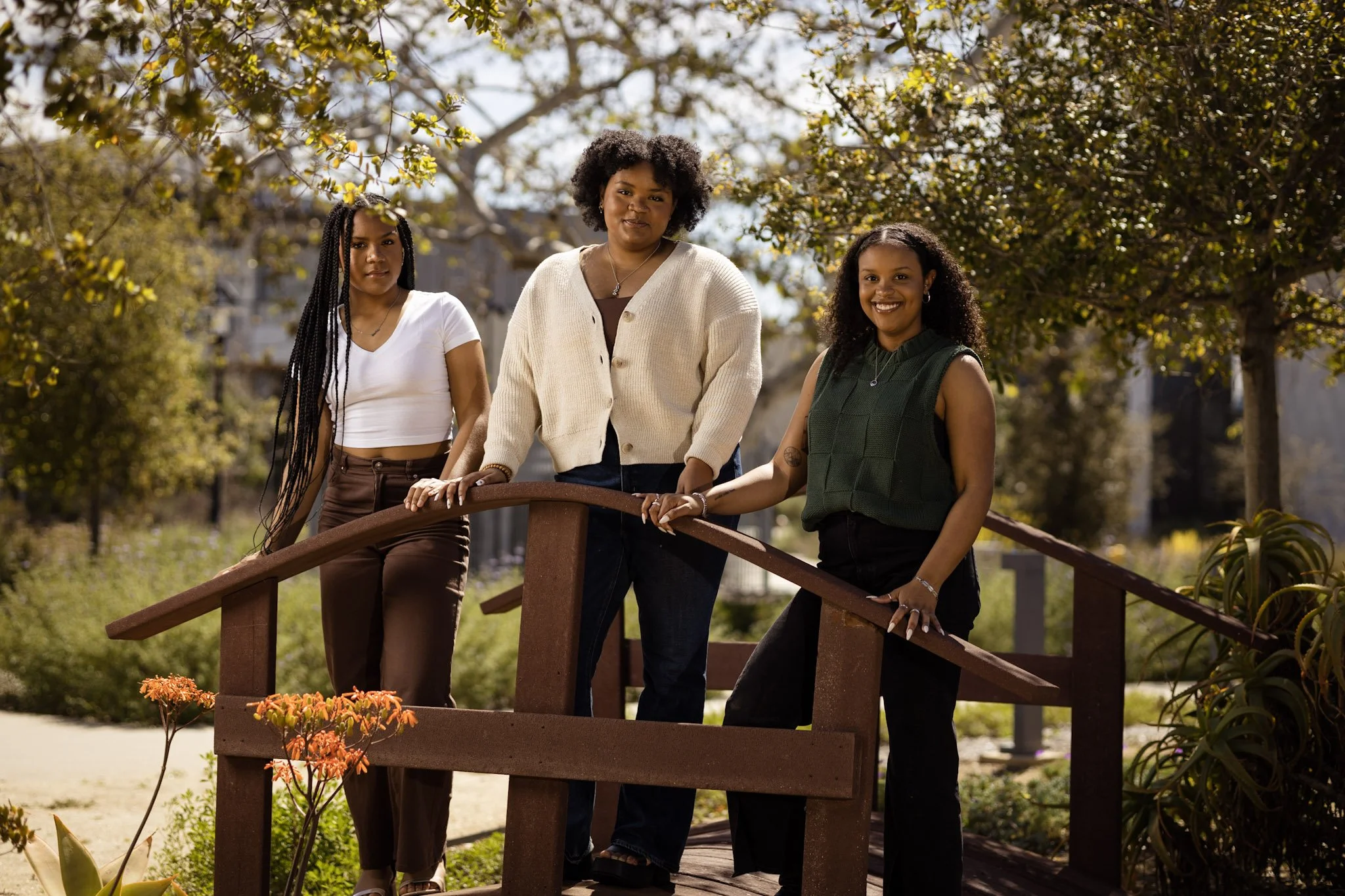 Three women standing on a small wooden bridge outdoors surrounded by trees and plants.