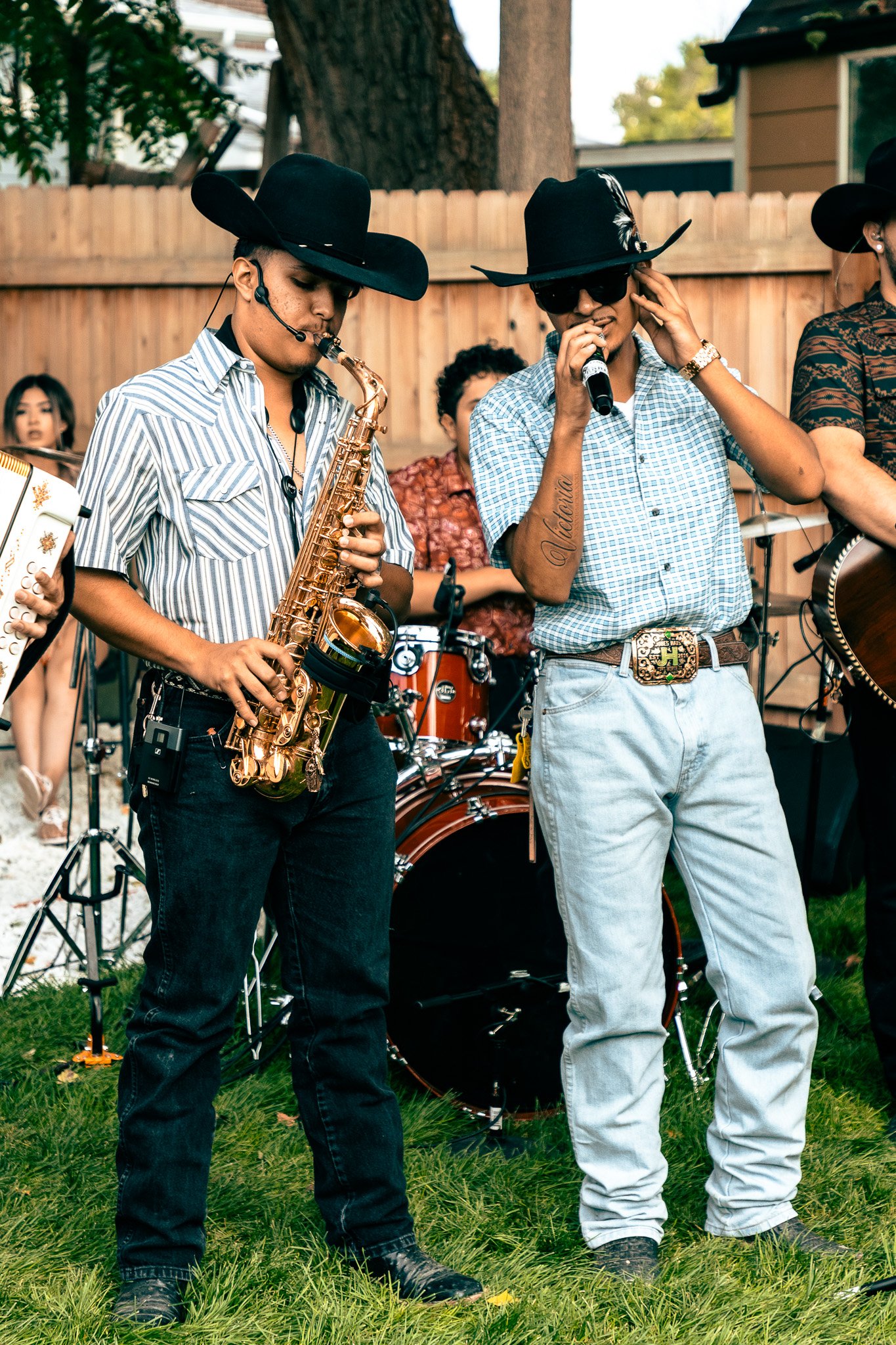 A group of musicians performing outdoors, with two men in cowboy hats in the foreground, one playing saxophone and the other singing into a microphone, surrounded by other band members and audience.
