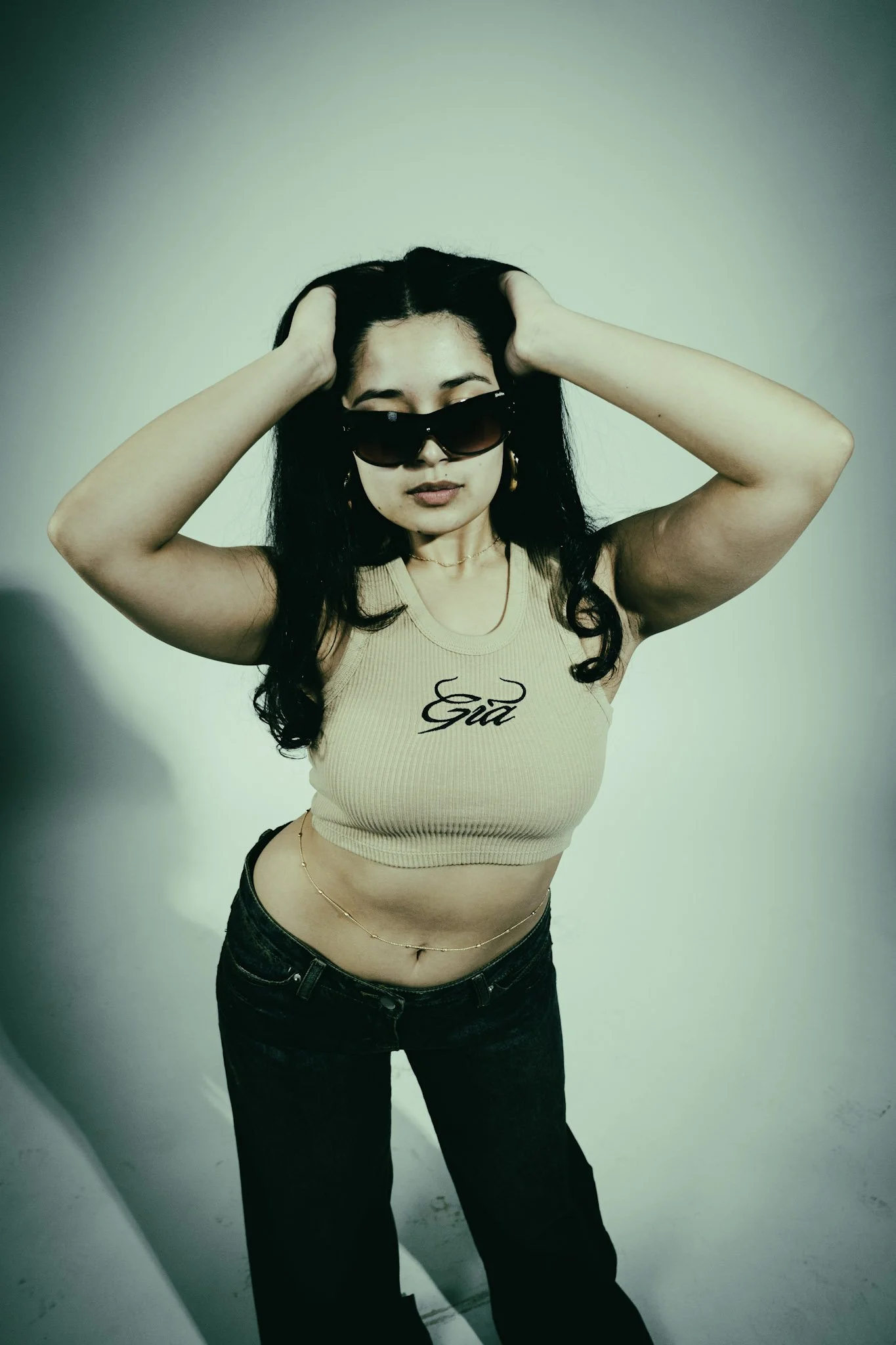 A woman with long black hair wearing large sunglasses, a sleeveless beige crop top with the word "Girl" written on it, dark jeans, and a gold chain belt, standing against a plain light green background.