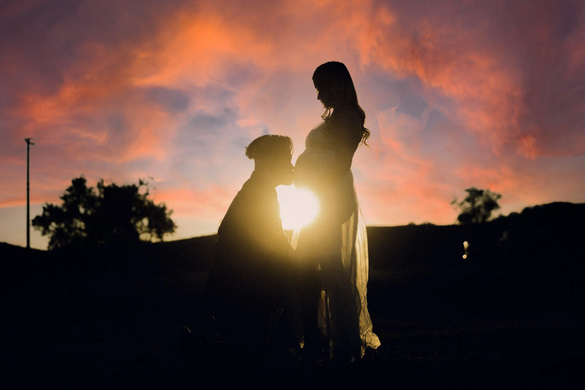 Silhouette of a pregnant woman standing and a person kneeling before her, with a sunset sky with colorful clouds in the background.