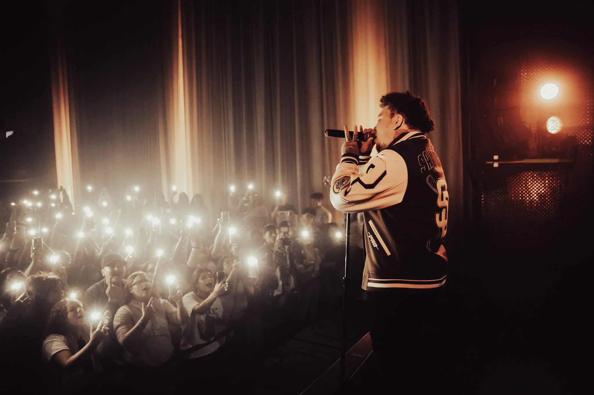 A male performer in a varsity jacket singing into a microphone on stage with an audience holding up their phones with flashlights on.