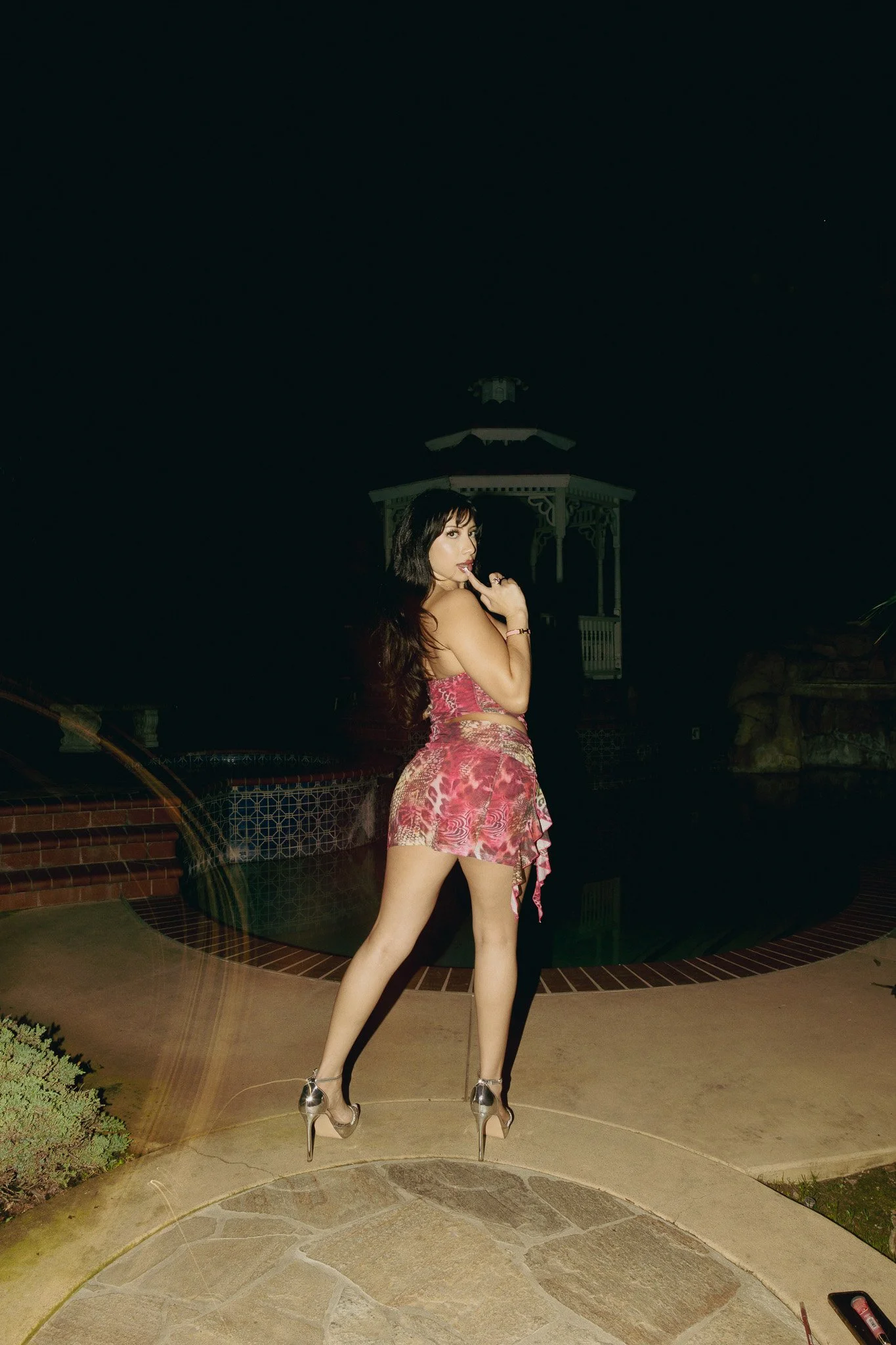A woman in a pink floral dress and high heels standing outdoors at night near a pool, with a dark sky in the background.