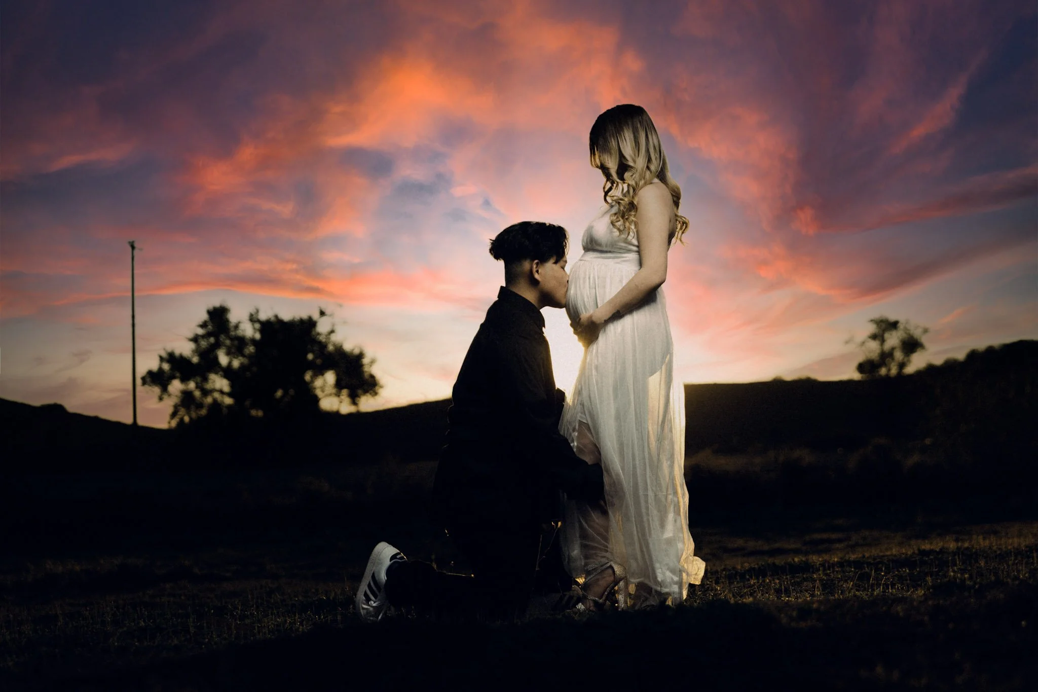 A man kneeling and kissing a pregnant woman’s belly outdoors at sunset, with a colorful sky and silhouettes of distant trees and hills.