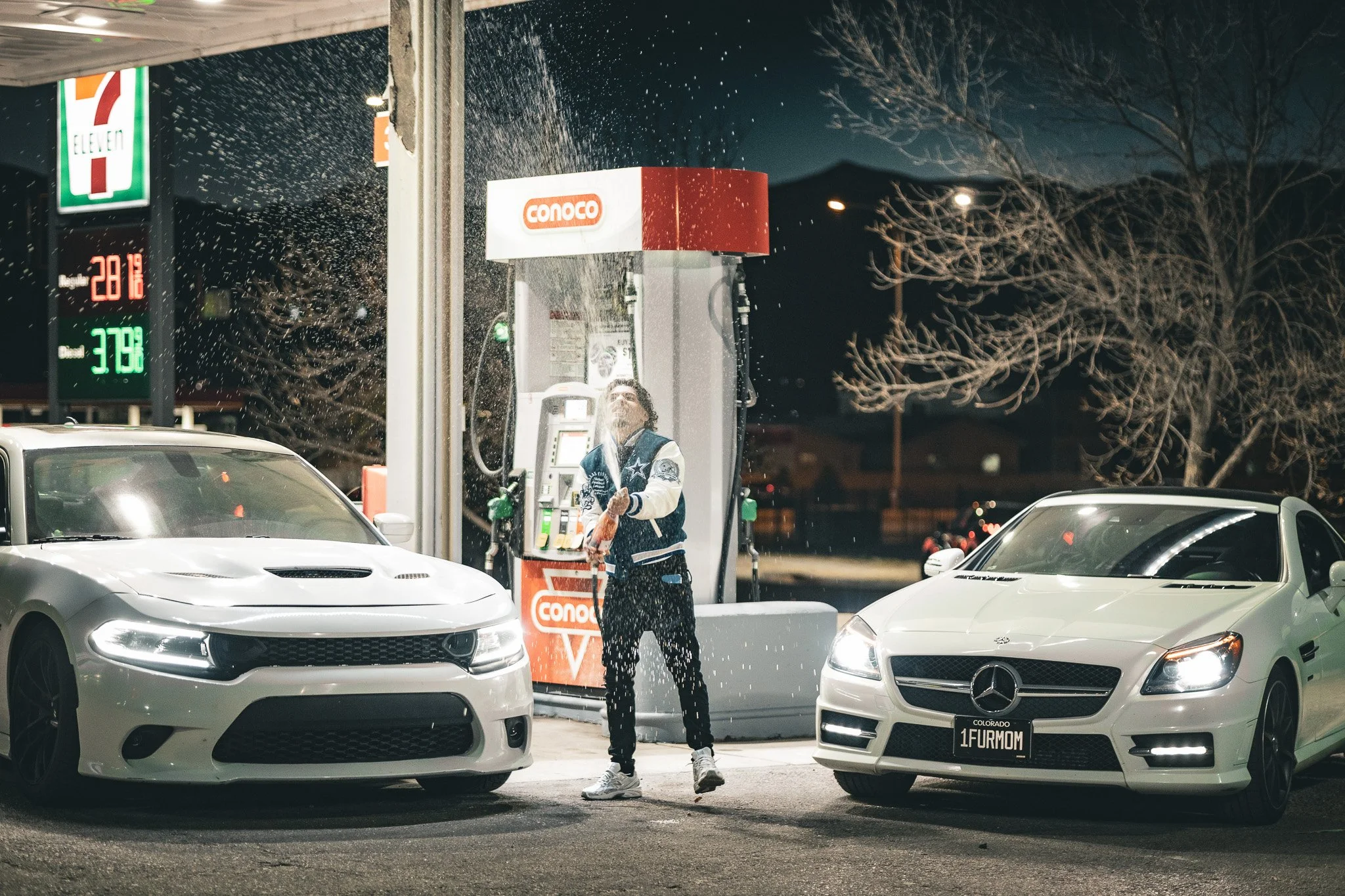 A person in a Dallas Cowboys jacket is washing a white car at a gas station during nighttime. The gas station has a Conoco sign and fuel pumps, with the person standing between two white cars, one on each side. A 7-Eleven sign and digital fuel prices