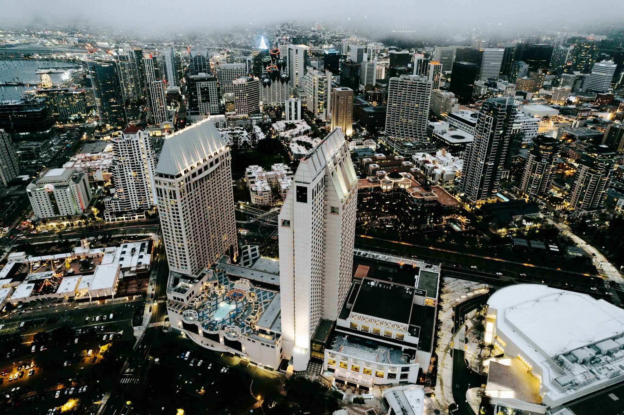 Aerial view of downtown San Diego at dusk, featuring tall skyscrapers, a hotel with a rooftop pool, and city lights beginning to illuminate the skyline.