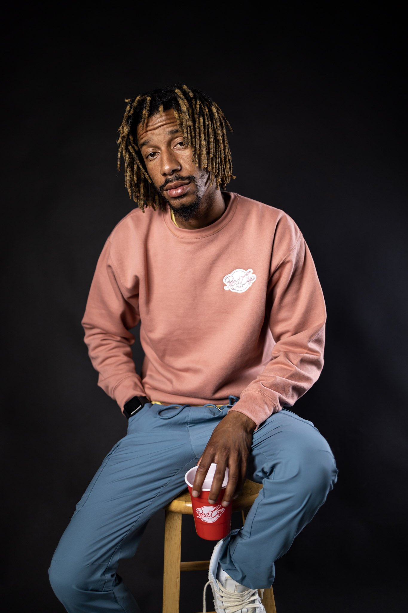 A young man with dreadlocks seated on a wooden stool against a black background, holding a red cup in his right hand, wearing a pink sweatshirt, blue pants, and white sneakers.