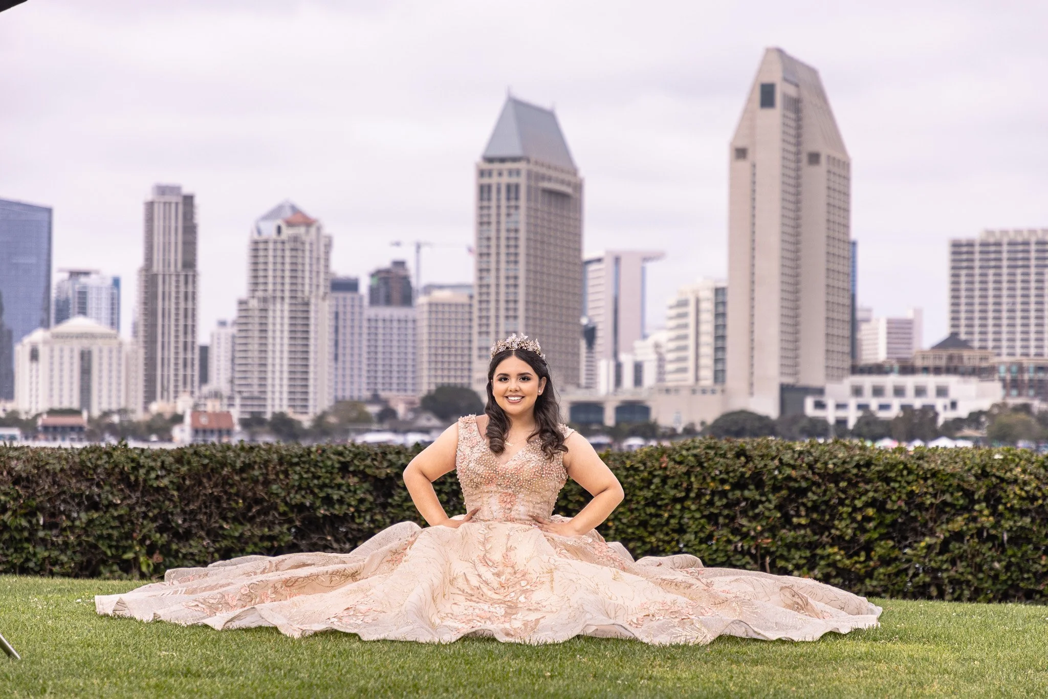 A woman in a light pink, ornate gown with a full skirt, sitting on green grass in front of a city skyline with tall buildings, wearing a tiara and smiling.