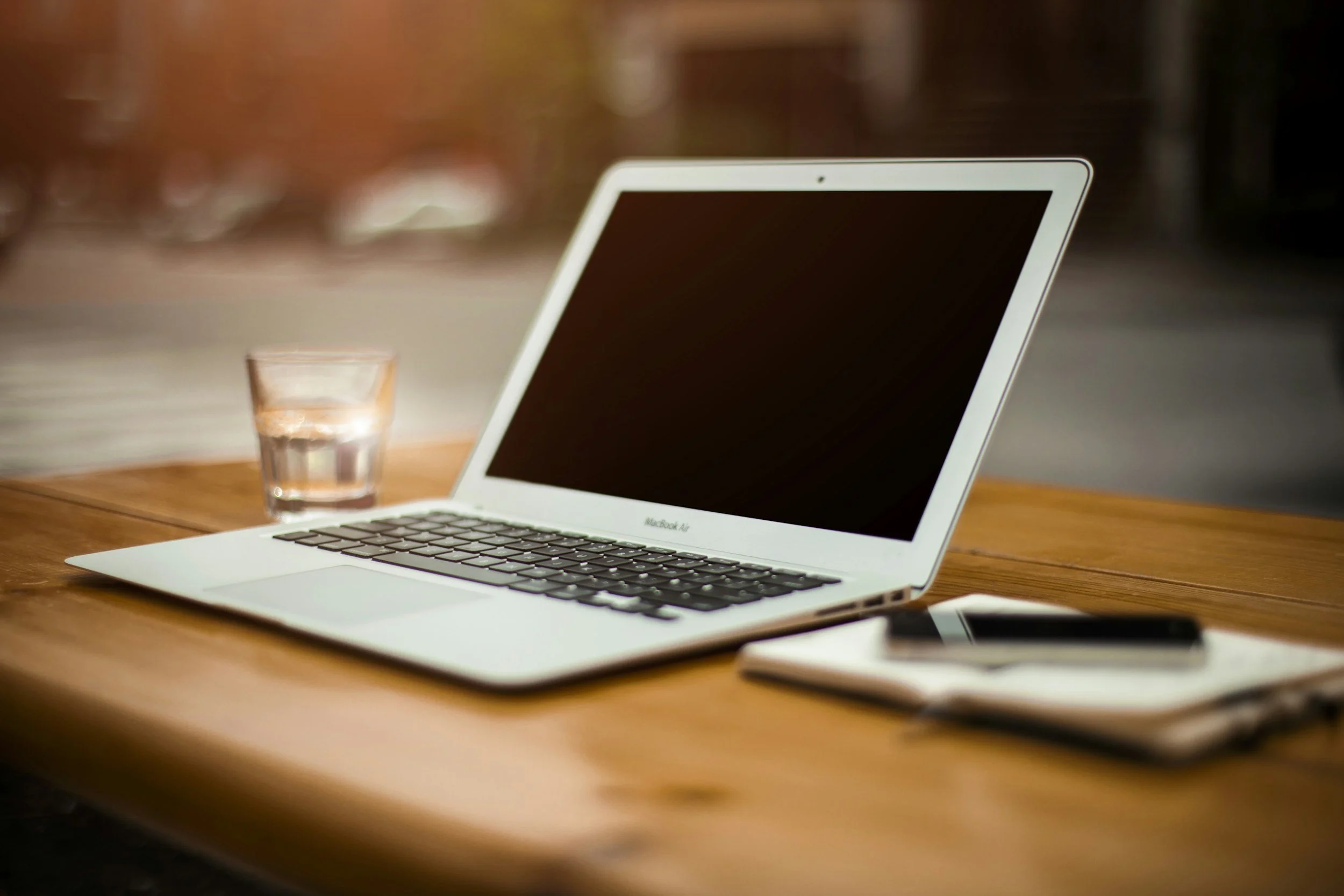Laptop on wooden desk with a glass of water, a notebook, and a smartphone.