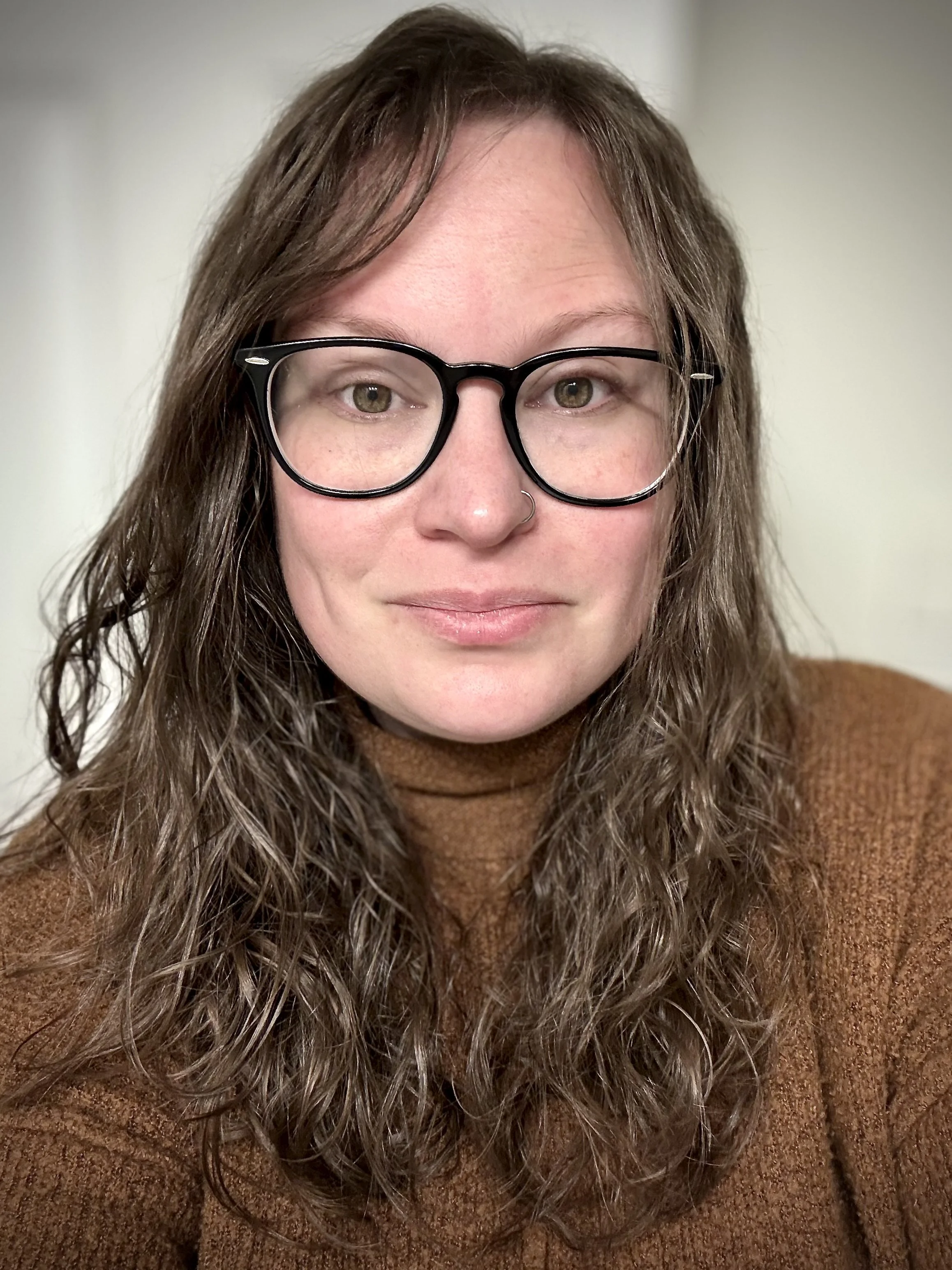 A woman with long, curly brown hair wearing black-rimmed glasses and a brown sweater, looking at the camera with a slight smile, in an indoor setting with a plain background.