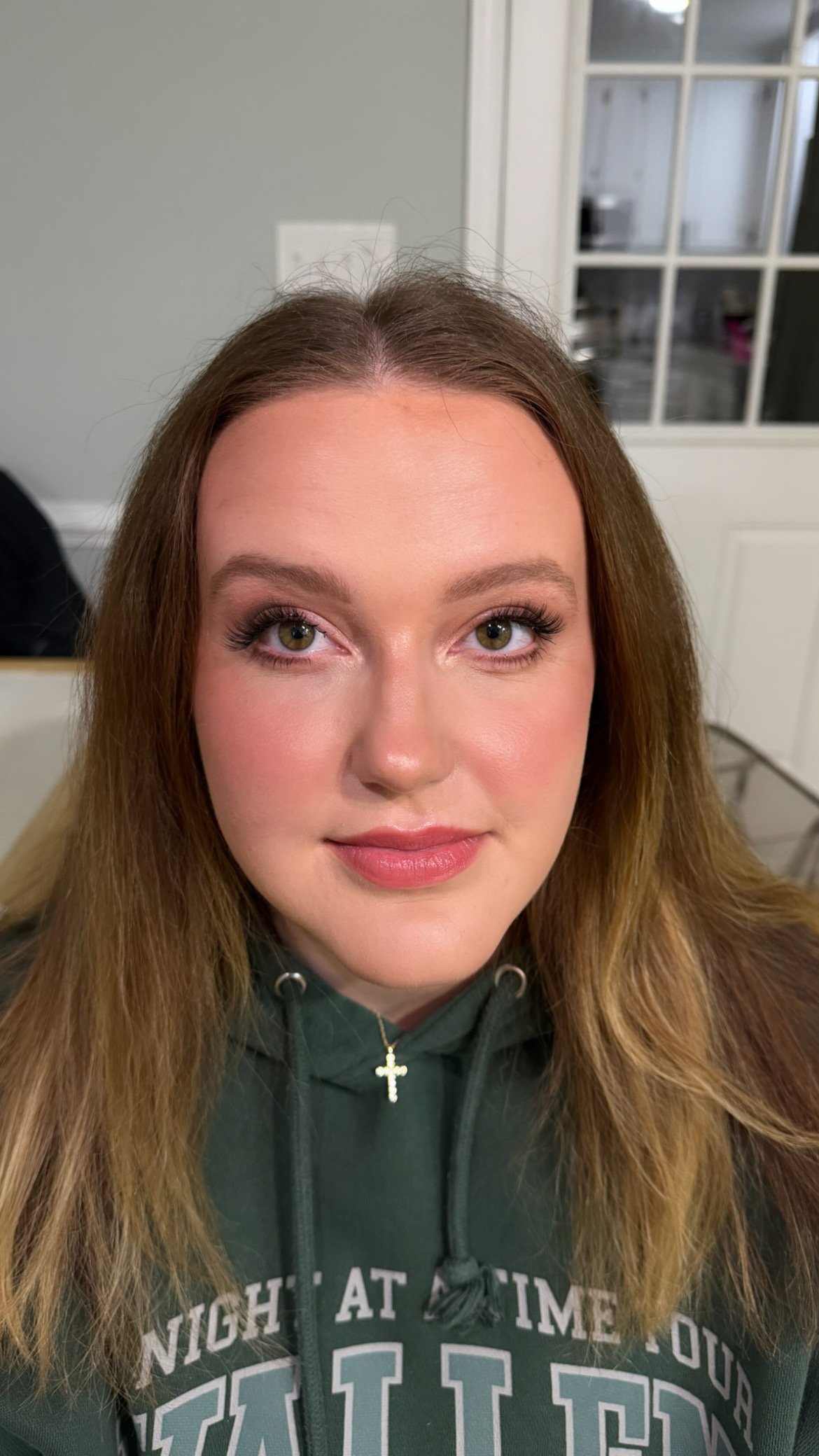Close-up of a young woman with long brown hair and green eyes, smiling softly, wearing a dark green hoodie with partial text and a gold cross necklace.