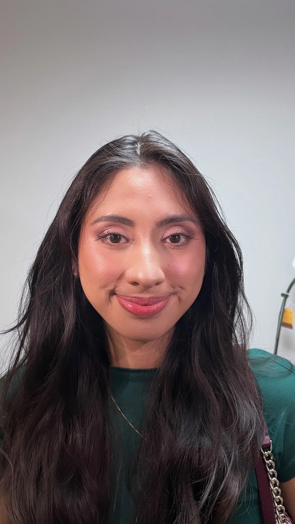 A portrait of a woman with long dark hair, pink makeup, and wearing a green top, standing indoors against a plain white wall.