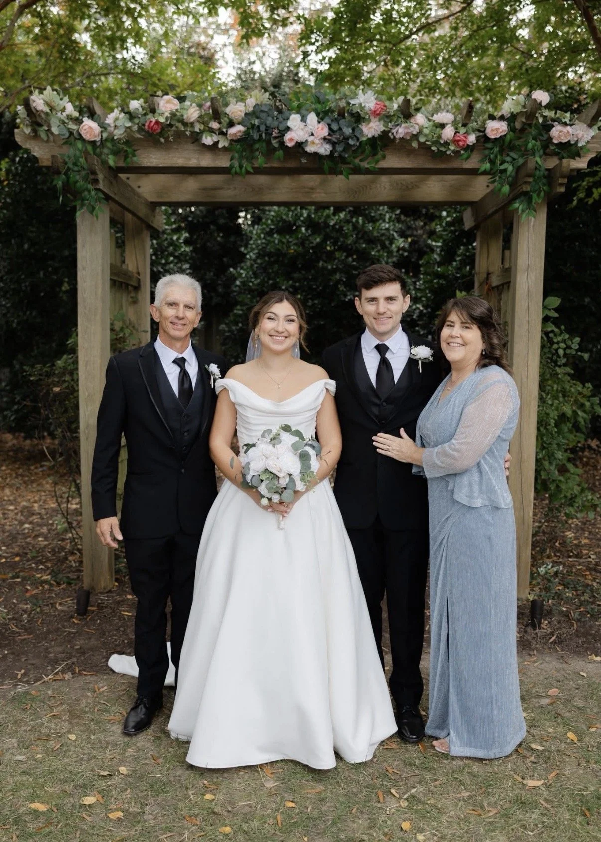 A wedding couple with family under a floral archway outdoors, with trees in the background. The bride in a white dress holds a bouquet, and the groom and family members are in formal attire.