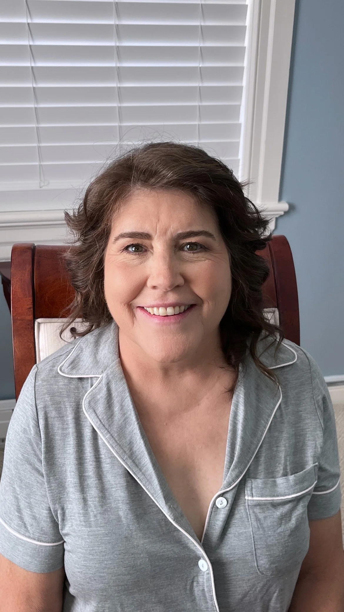 A woman with shoulder-length wavy brown hair, wearing gray pajamas with white piping, smiling and sitting in a wooden chair with a high back.