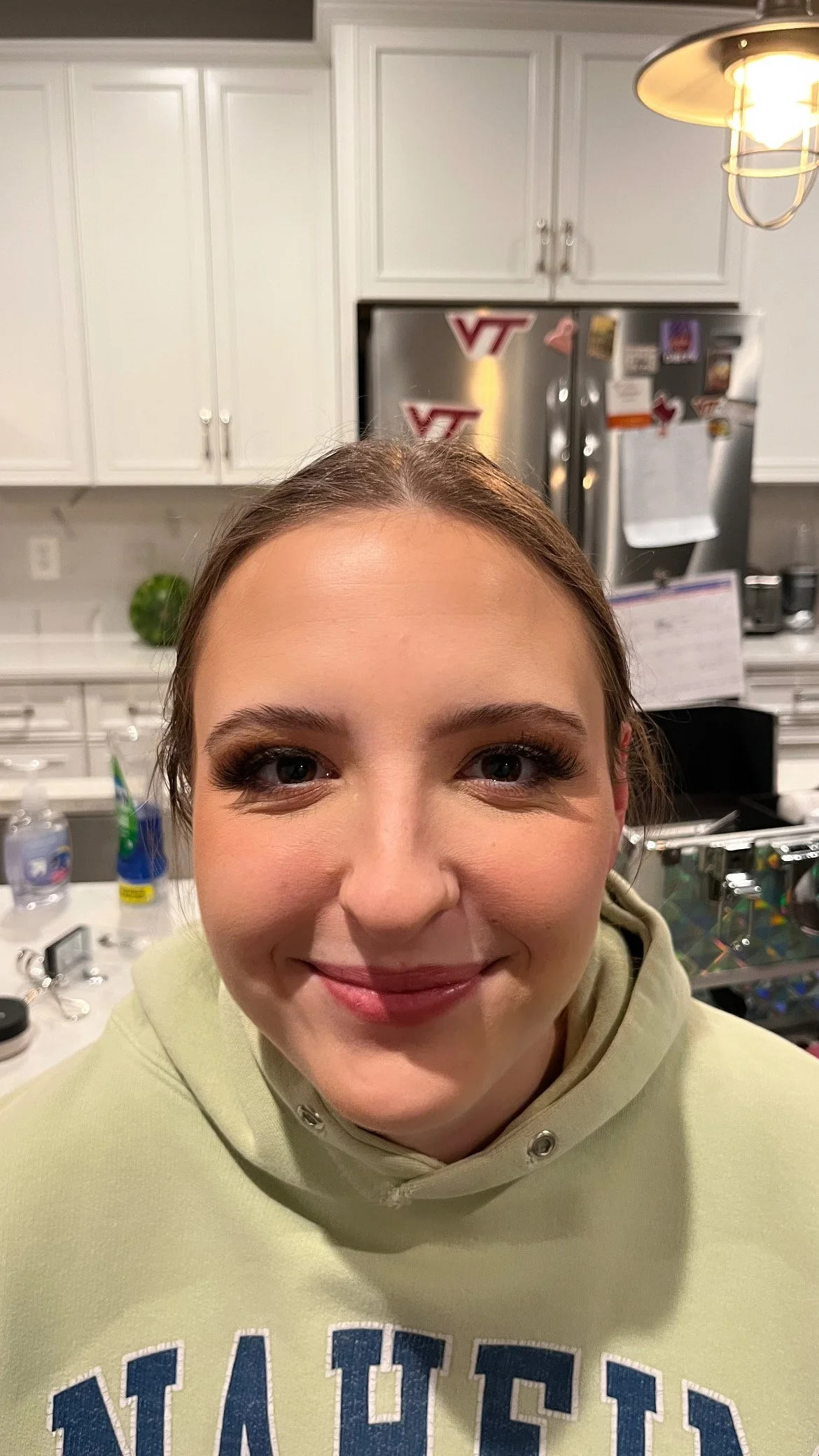 Close-up of a smiling woman with brown hair, wearing a light-colored hoodie, in a kitchen with white cabinets and a stainless steel refrigerator in the background.