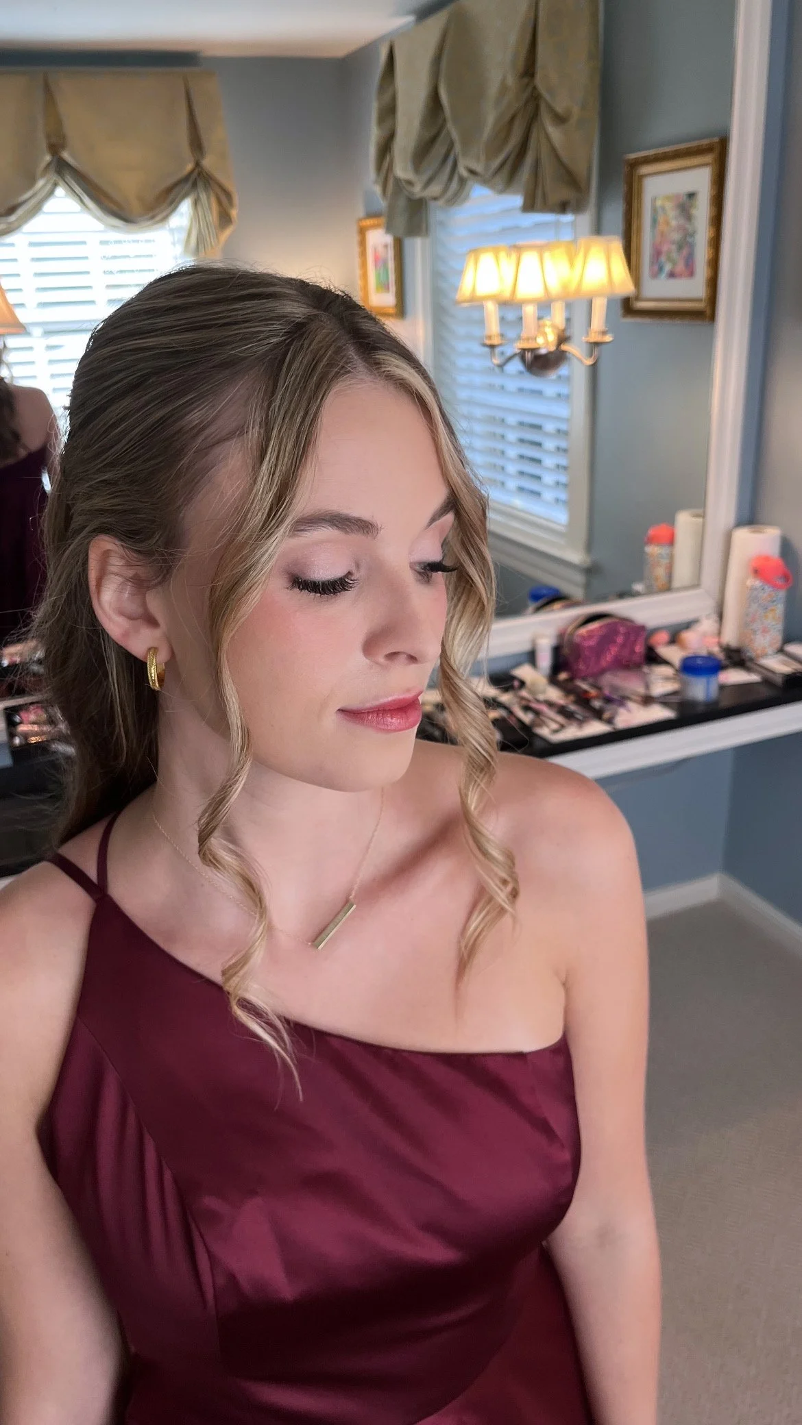 A young woman with light skin and blonde hair styled in loose curls, wearing a burgundy dress, gold earrings, and a gold necklace, sitting in a room with a vanity and makeup items in the background.