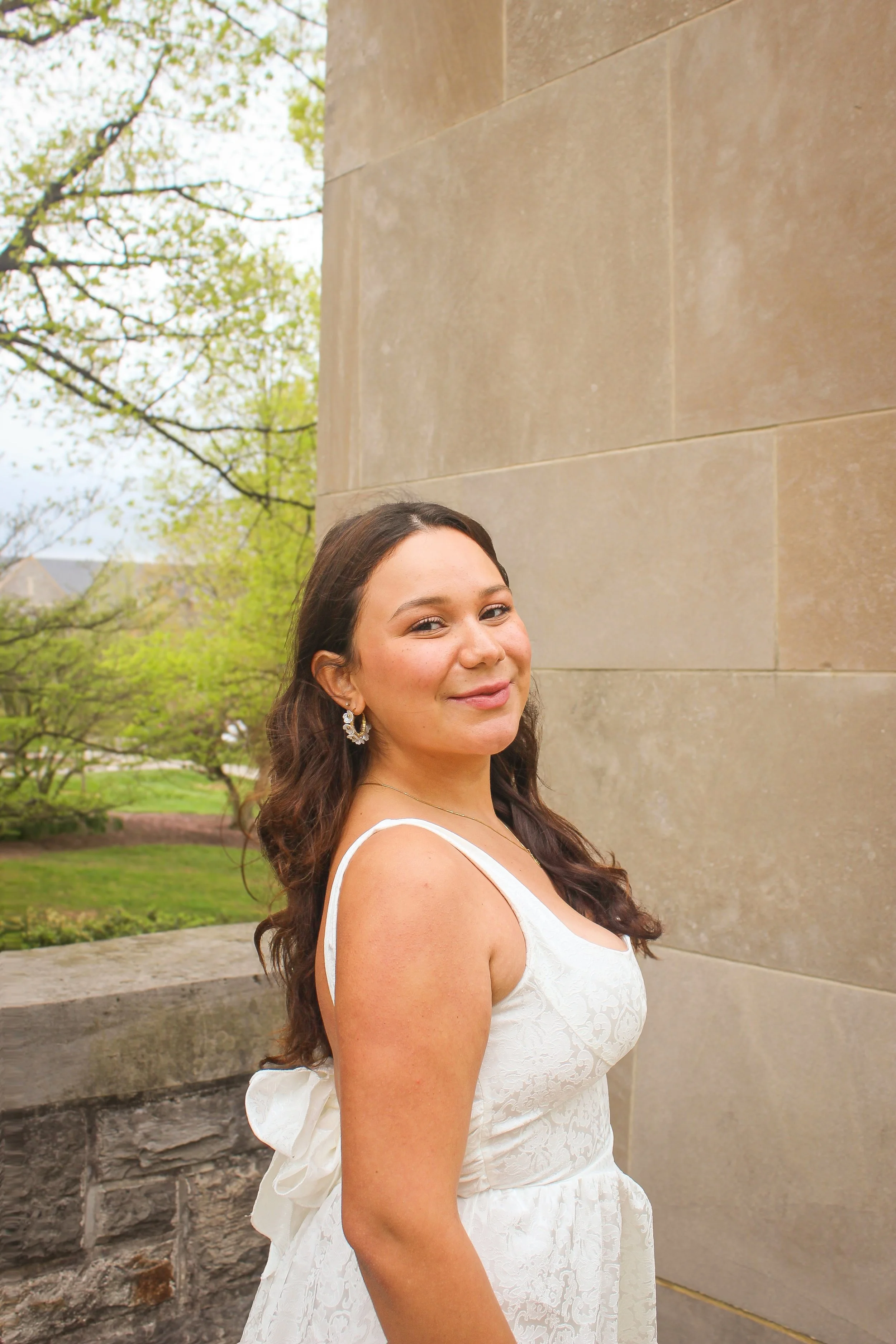 A woman in a white dress standing outdoors next to a stone building with trees in the background.