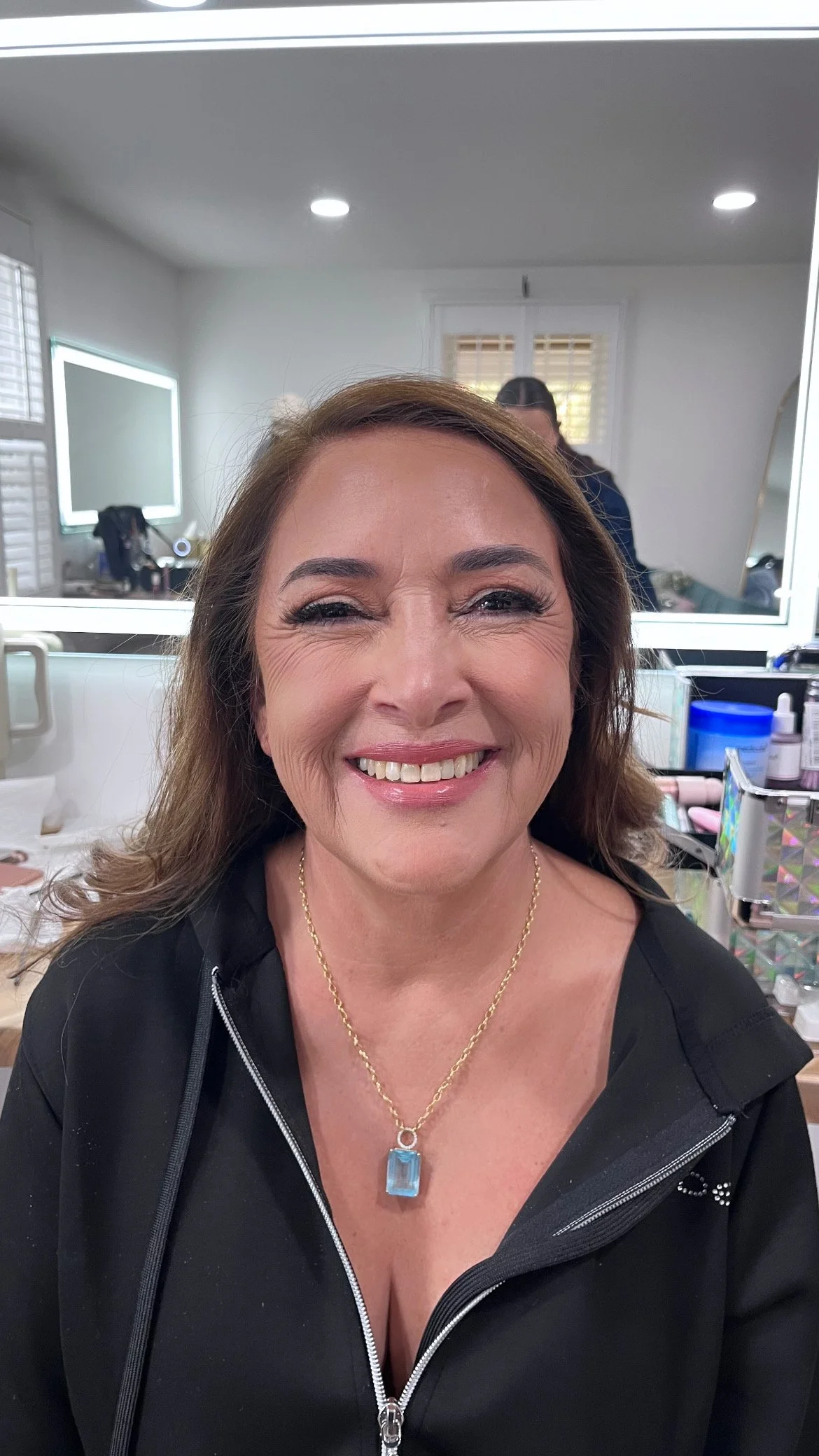 A woman smiling confidently, wearing a black jacket and a gold chain with a blue gemstone pendant, in a makeup or hair styling room.
