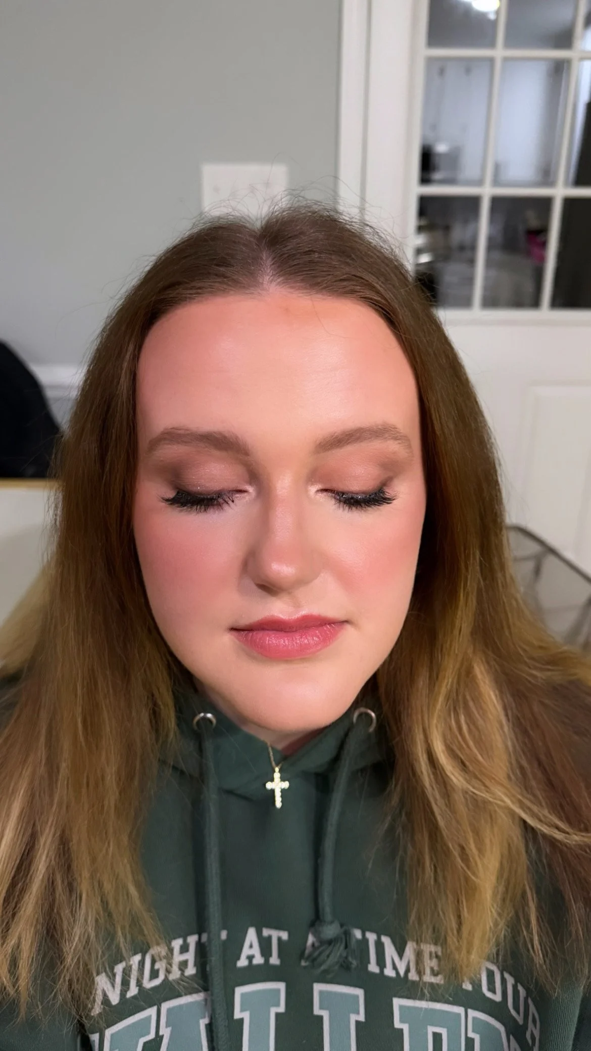 A young woman with closed eyes and makeup, wearing a green hoodie with white text and a gold cross necklace, in an indoor setting with a glass door and light gray wall in the background.