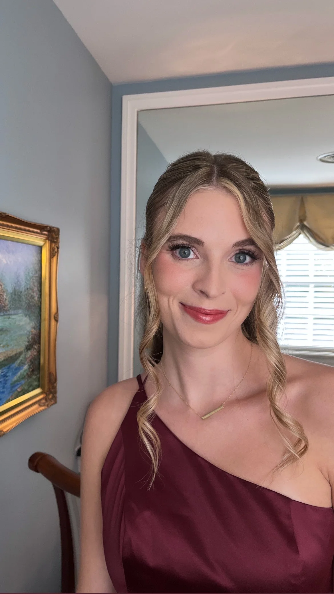 A young woman with blonde, curly hair in a half-up style, wearing a burgundy dress and a thin gold necklace, smiling in a well-lit room with blue walls, a framed painting, a window with blinds and a gold valance.