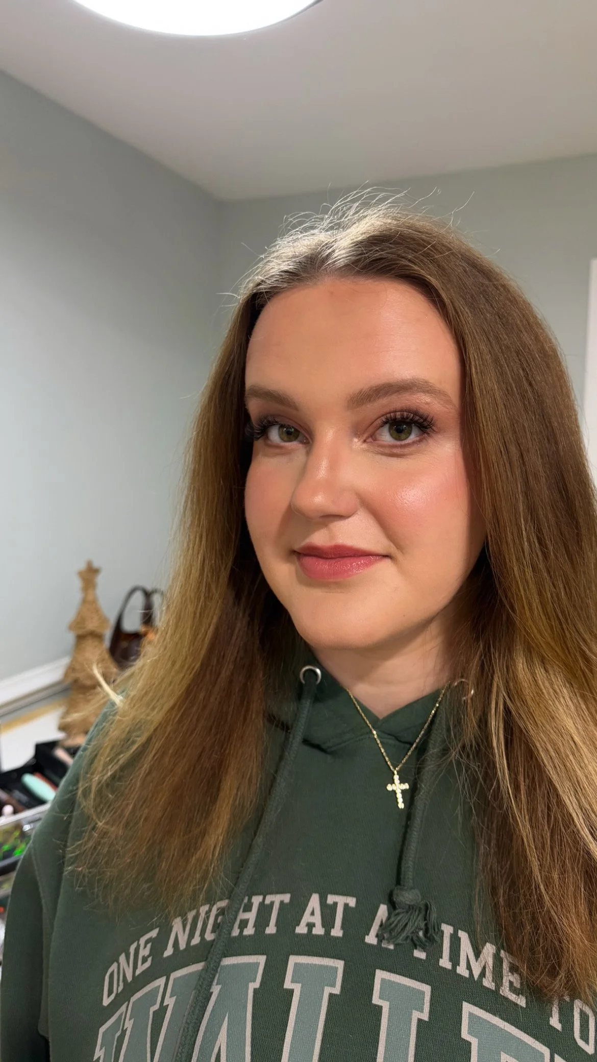 A woman with light skin and straight, shoulder-length brown hair, wearing makeup and a dark green hoodie with text, standing in a room with a light-colored wall and some decorative items in the background.