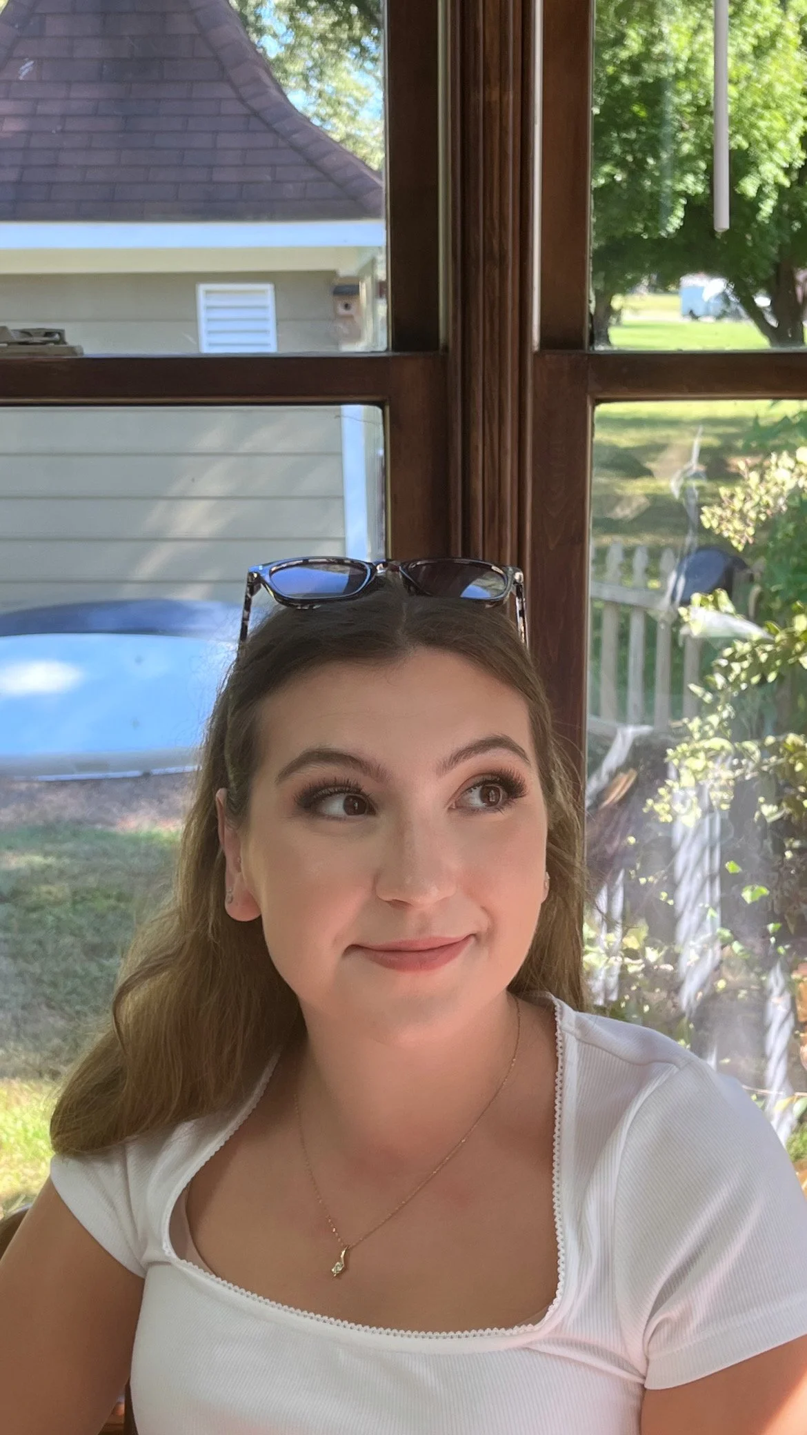 A young woman with brown hair, wearing a white shirt and sunglasses on her head, looking to her right inside a sunlit room with large windows. Outside, trees, a fence, and part of a house are visible.