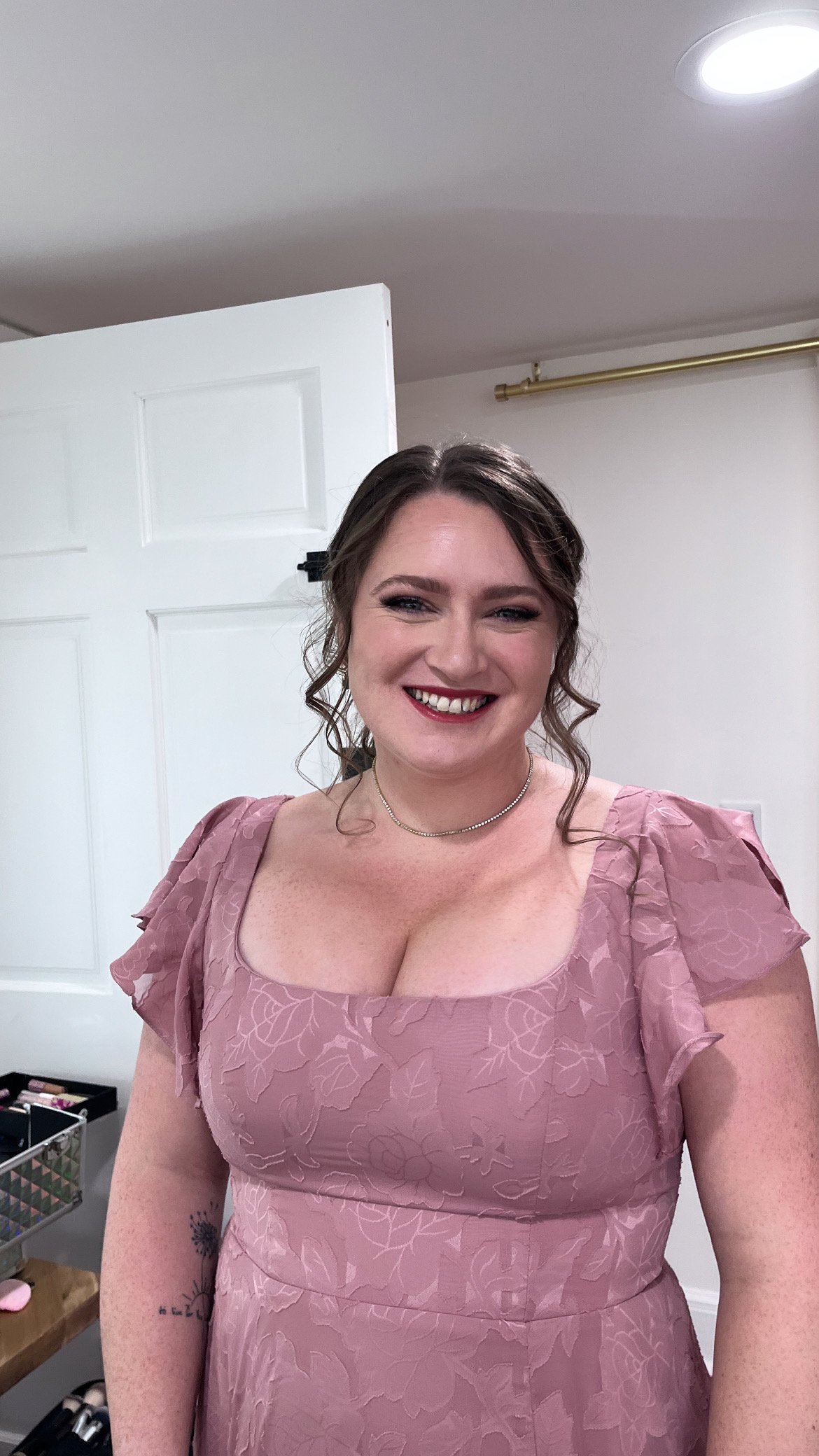 A woman with light skin, curly brown hair, and red lipstick, smiling in a pink dress with floral patterns, standing in a room with white walls and a white door.