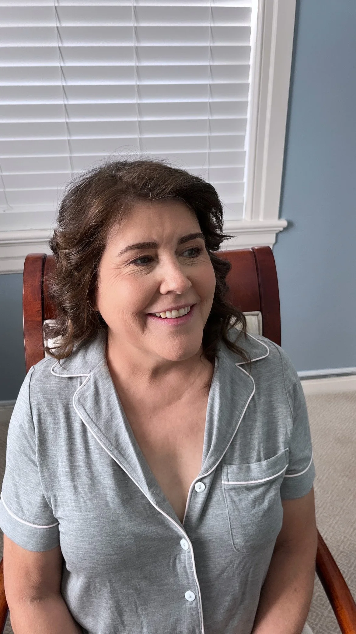 A smiling woman with shoulder-length brown hair sitting on a wooden chair in front of a window with white blinds, dressed in gray pajamas with white piping.
